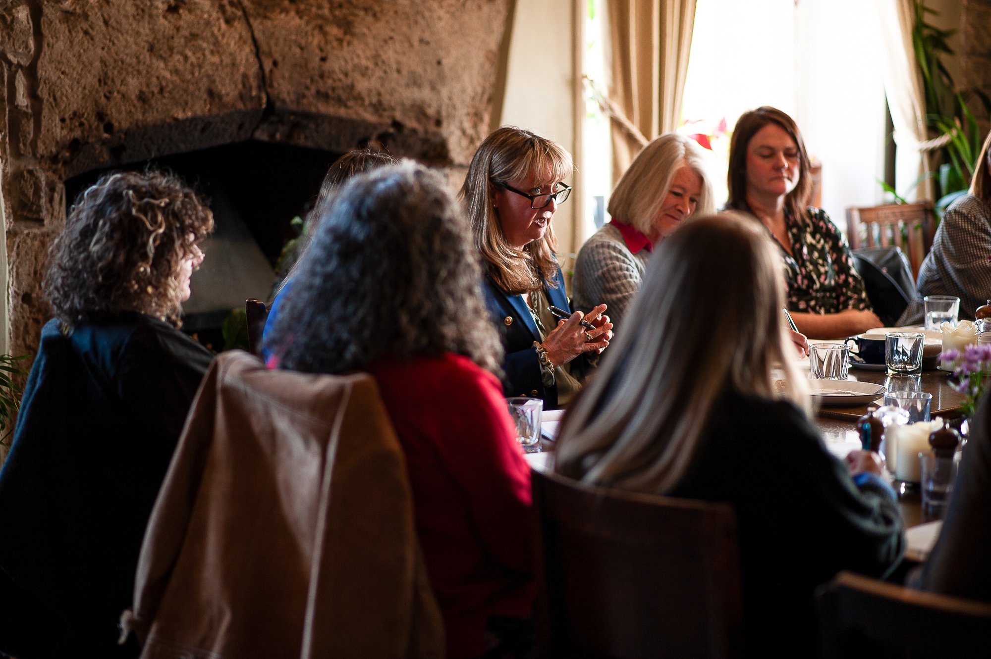Group of women sitting around a dining table in a cozy indoor setting, some taking notes, with natural light coming through a window.