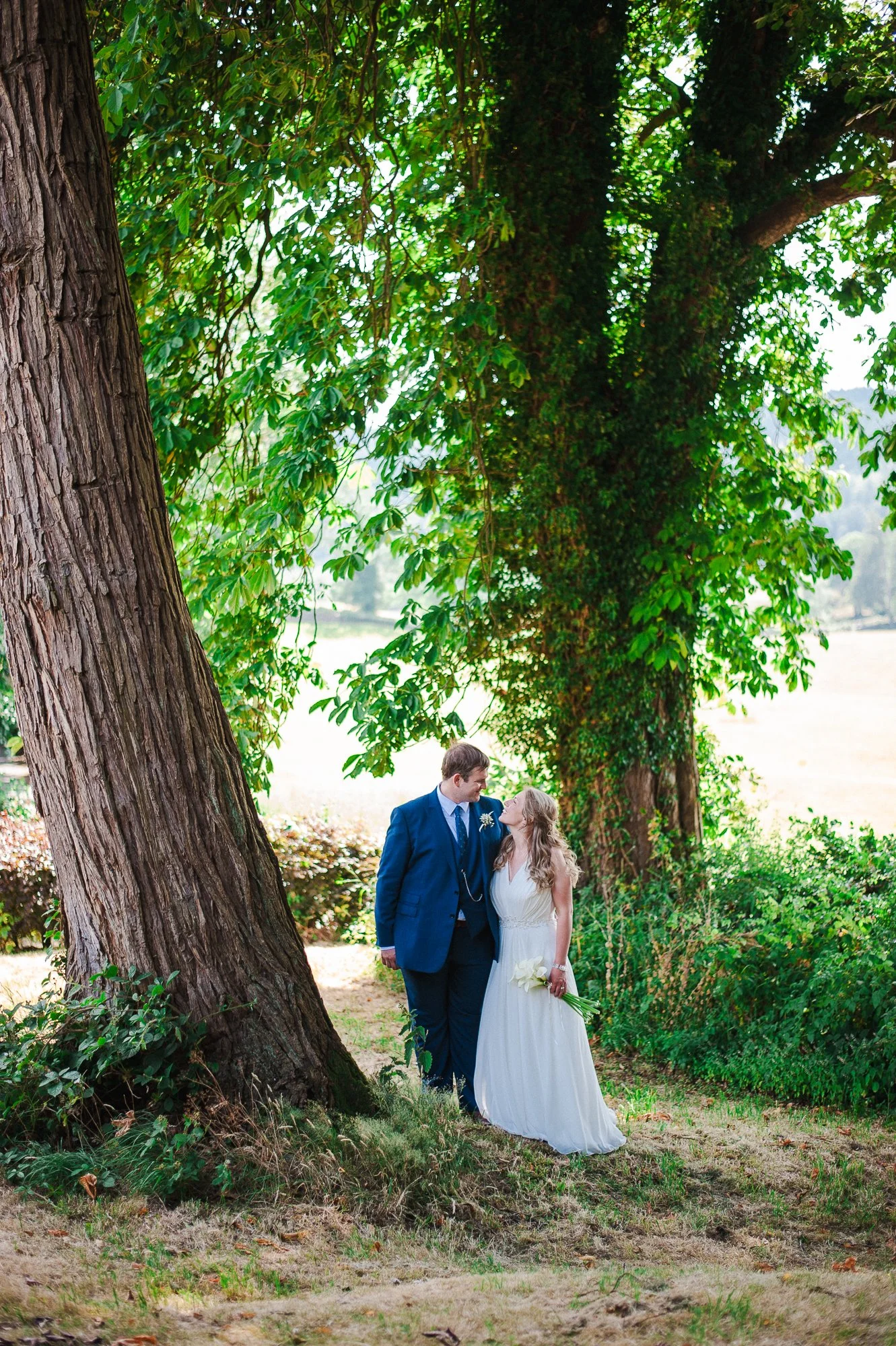 A newlywed couple stands beneath large trees in a lush outdoor setting, gazing at each other lovingly, the bride holding a bouquet of white flowers.