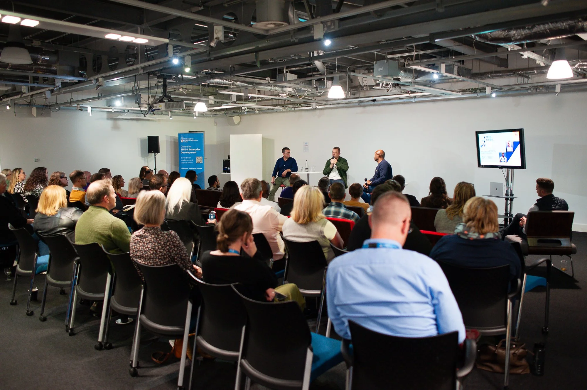 A conference room with an audience listening to three speakers on a stage, with a screen displaying a presentation on the right side.