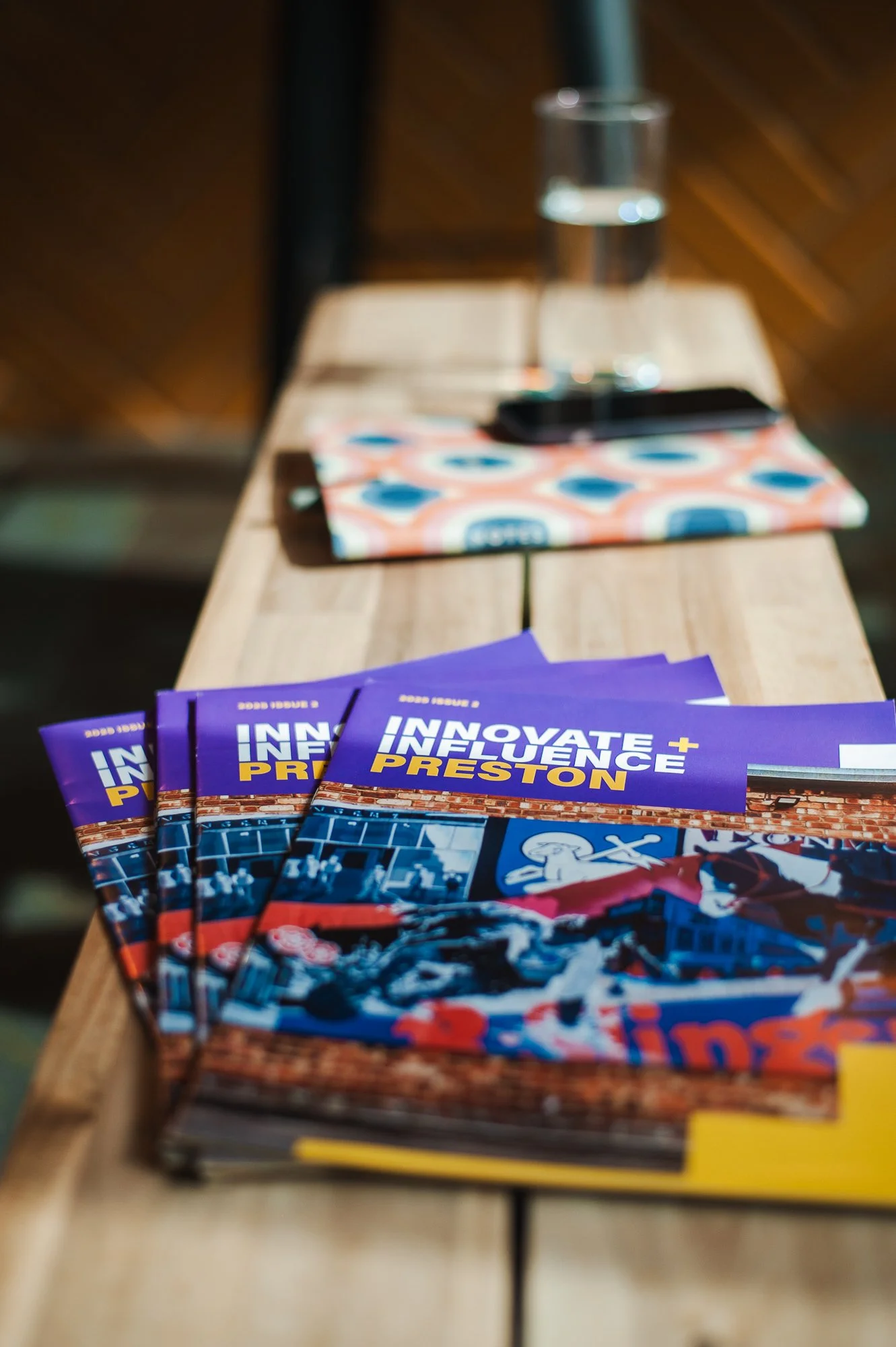 A stack of magazines titled 'Innovate + Influence Preston' on a wooden table, with a glass of water, colorful napkin, and smartphone in the background.