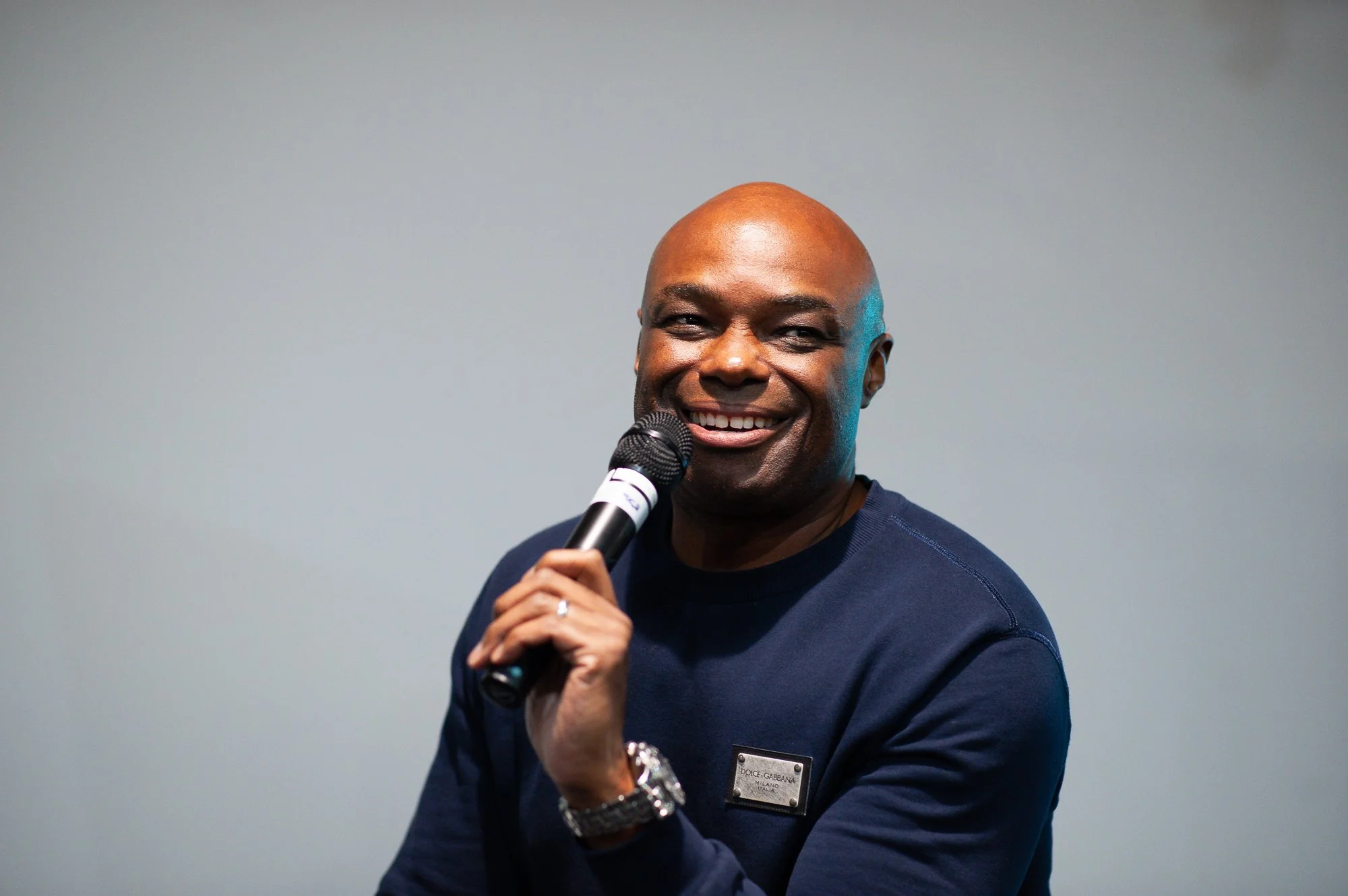 A smiling man holding a microphone, wearing a navy blue sweater with a name tag, standing against a neutral background.