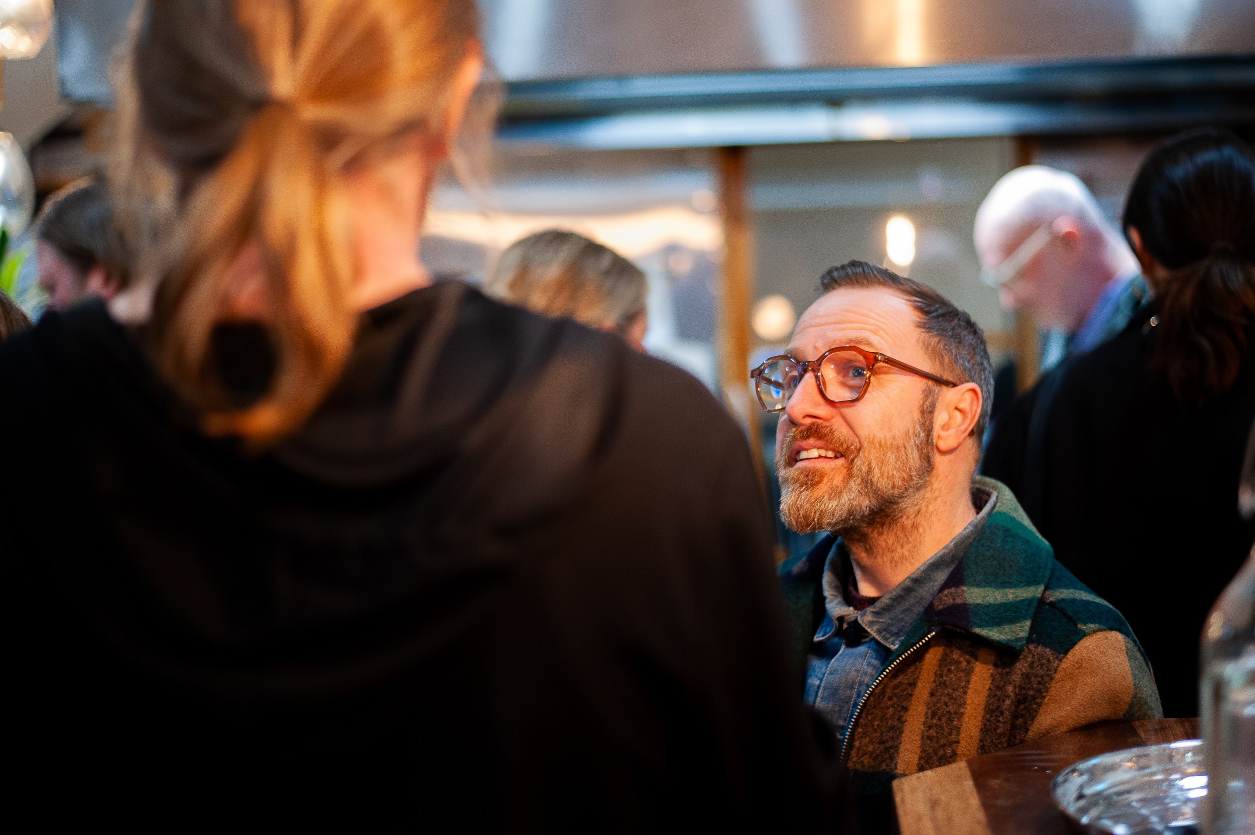 Man wearing glasses and a plaid jacket talking to a person with long hair in a social setting.