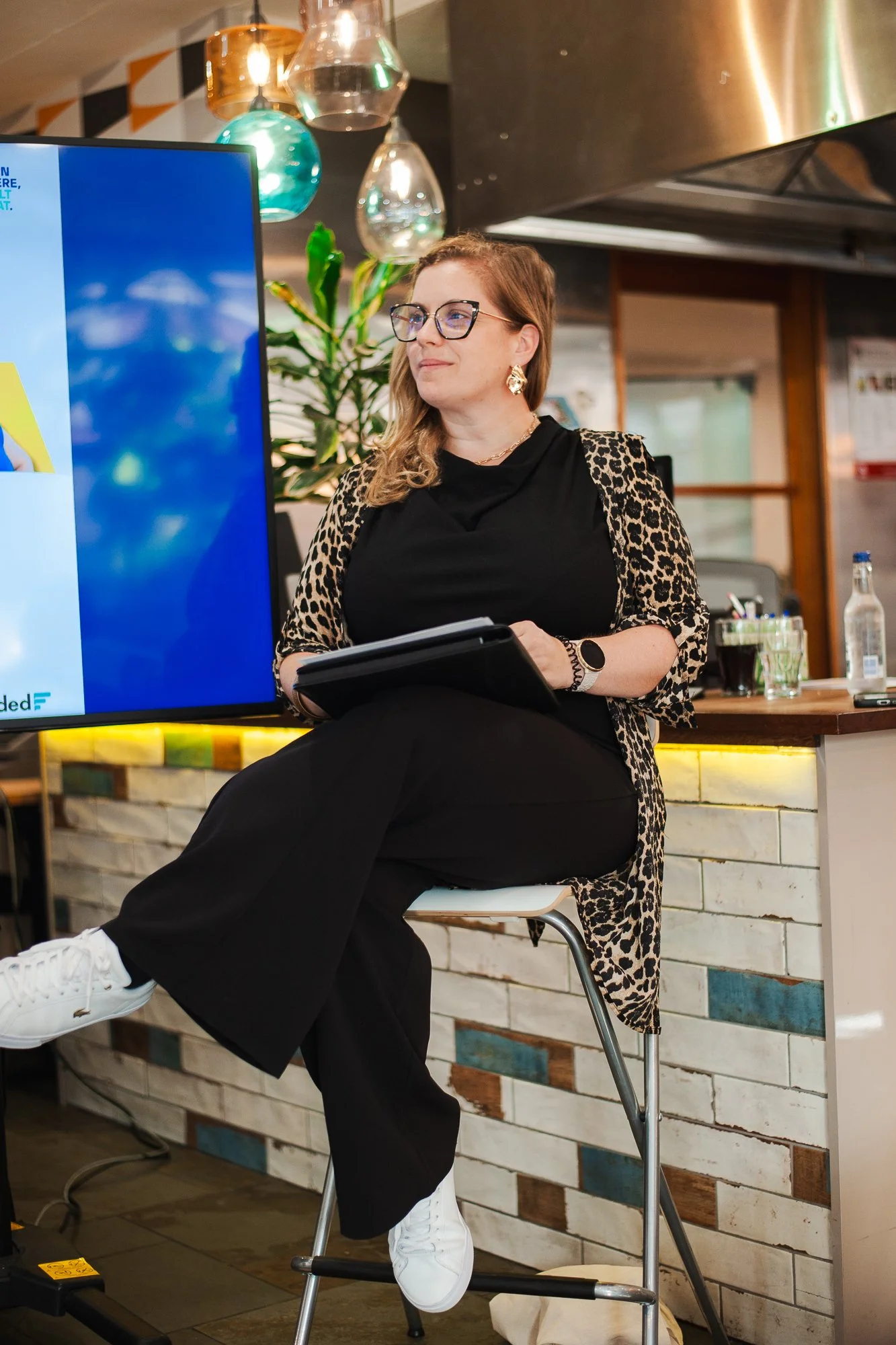 A woman with glasses and earrings sitting on a stool in a modern indoor setting, watching a presentation on a large screen. She's dressed in black pants, a black top, and a leopard print cardigan. There are decorative hanging glass lights and a bar c