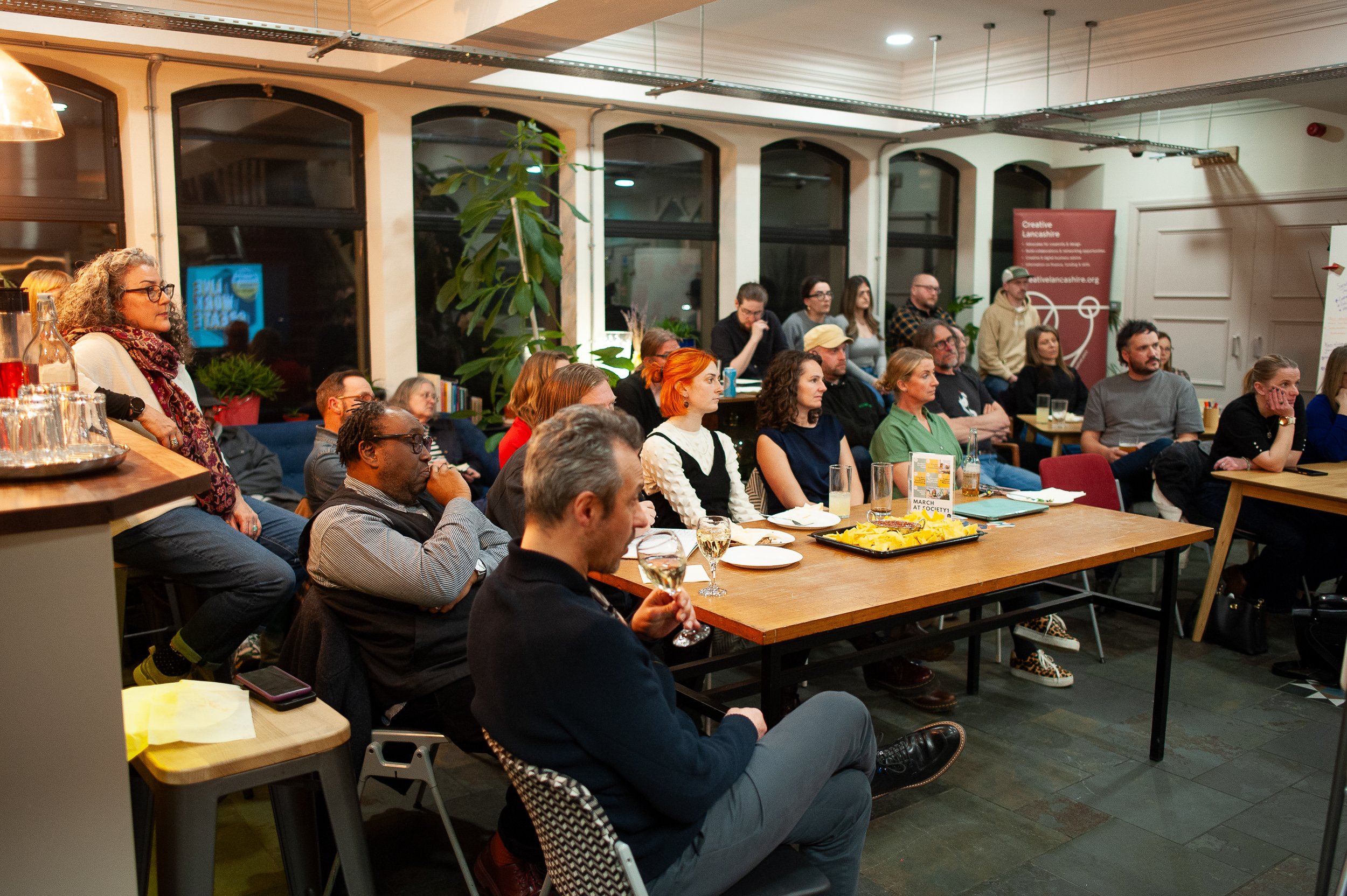 Group of people attending a presentation or discussion in a cozy room with tables, chairs, and plants.