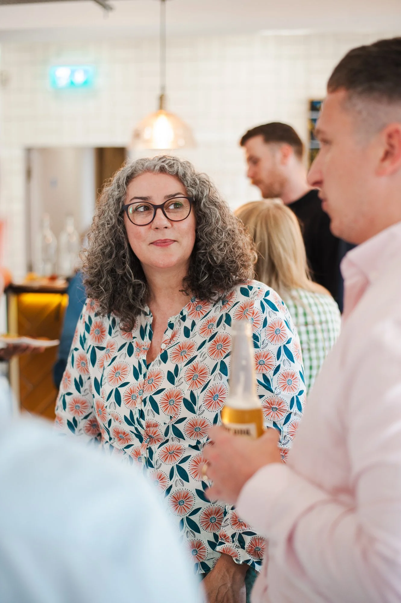 A woman with glasses and curly gray hair at a social gathering, wearing a floral blouse, engaging in conversation with a man holding a beer bottle.