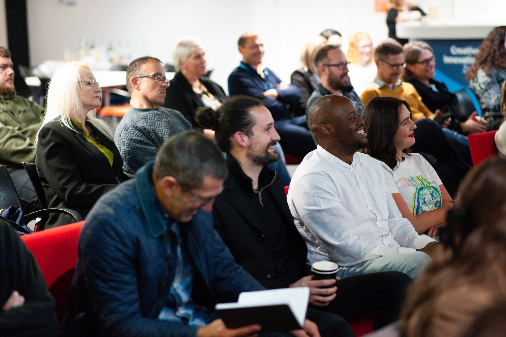 Audience members attending a conference or seminar, sitting in rows and smiling, some taking notes or holding coffee cups.