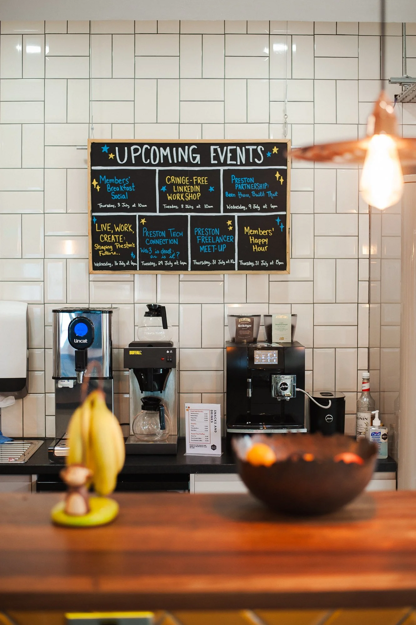 A blackboard menu listing upcoming events in a café or community space, with some hot beverage machines and a bowl of fruit on the counter below.