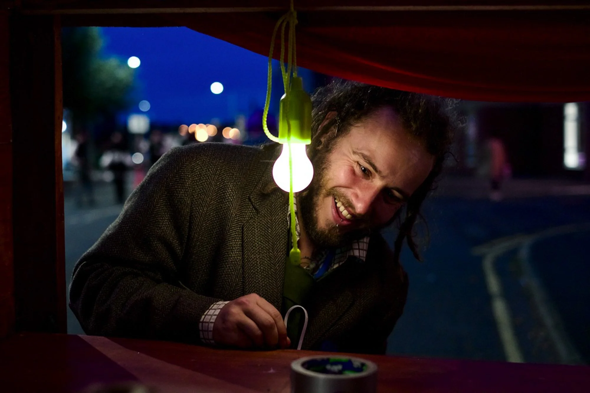 A man with long hair and a beard smiling, leaning inside a small booth or stall at night, with a lit green and white light bulb hanging nearby. The background shows a blurry outdoor setting with dark sky and distant city lights.