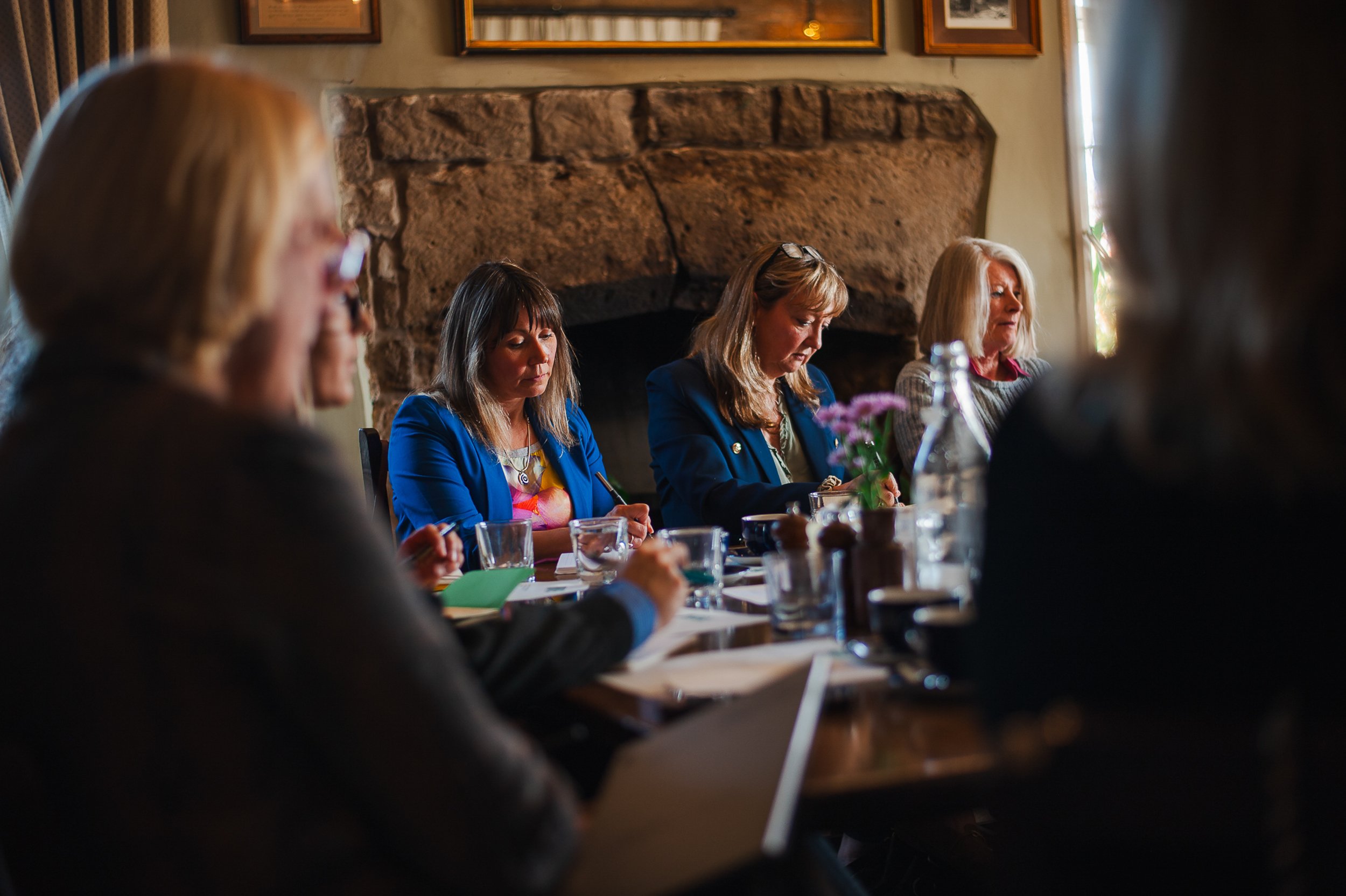 Group of people sitting around a table in a cozy room with stone fireplace, engaged in discussion or meeting.