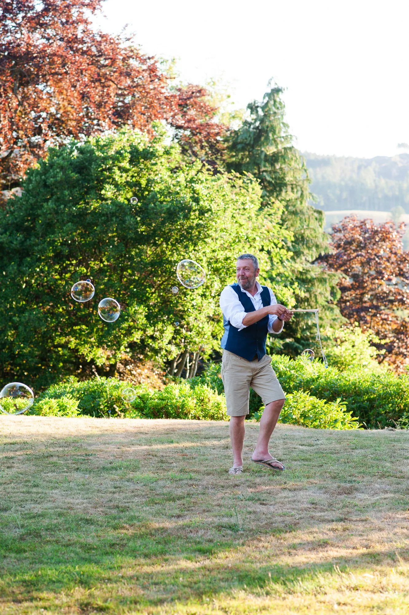 A man with a beard, wearing beige shorts, a white shirt, and a blue vest, playing with soap bubbles in a lush green garden during daytime.