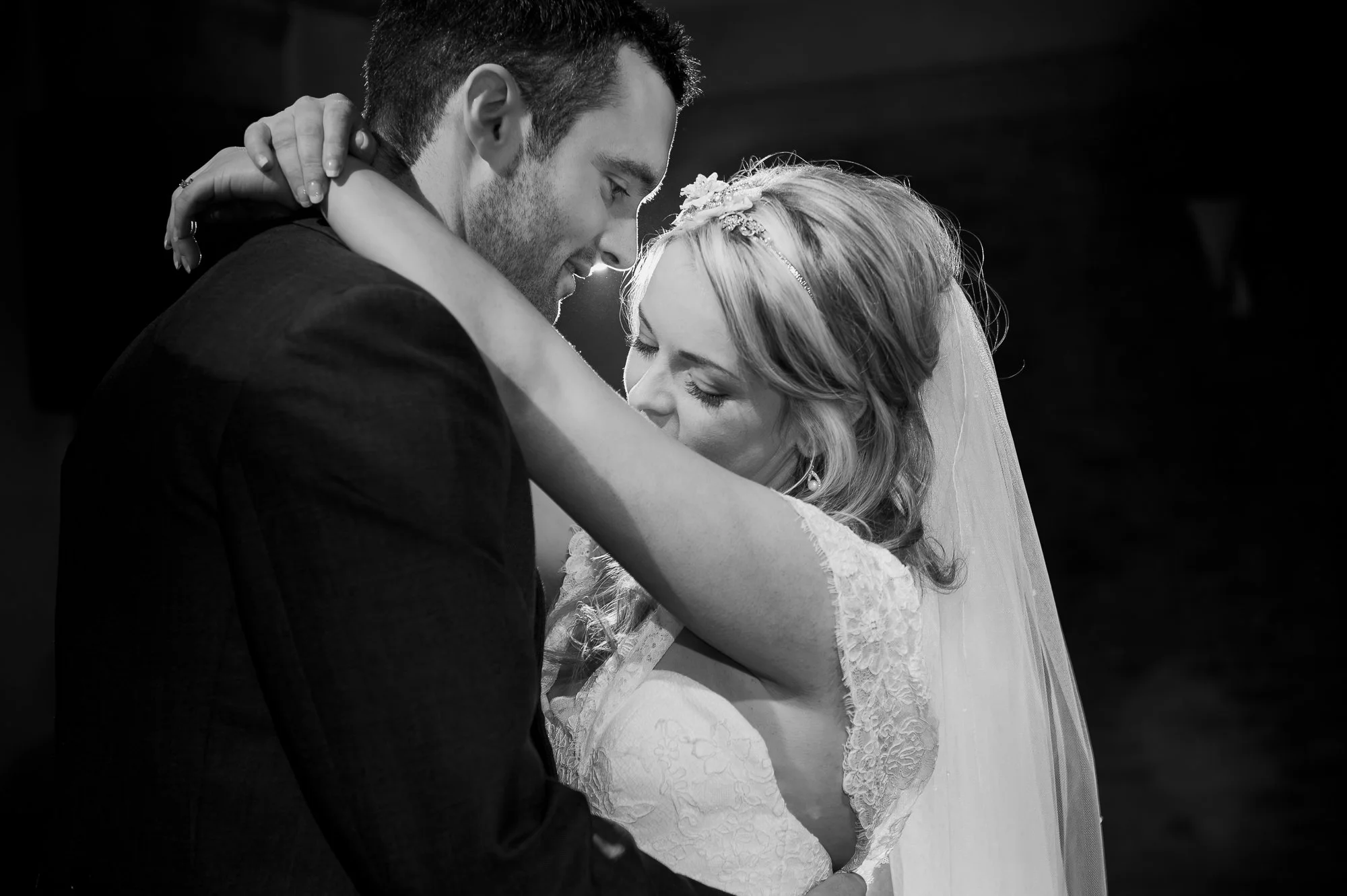 A black and white photograph of a wedding couple embracing, with the groom holding the bride close and both eyes closed, smiling softly.