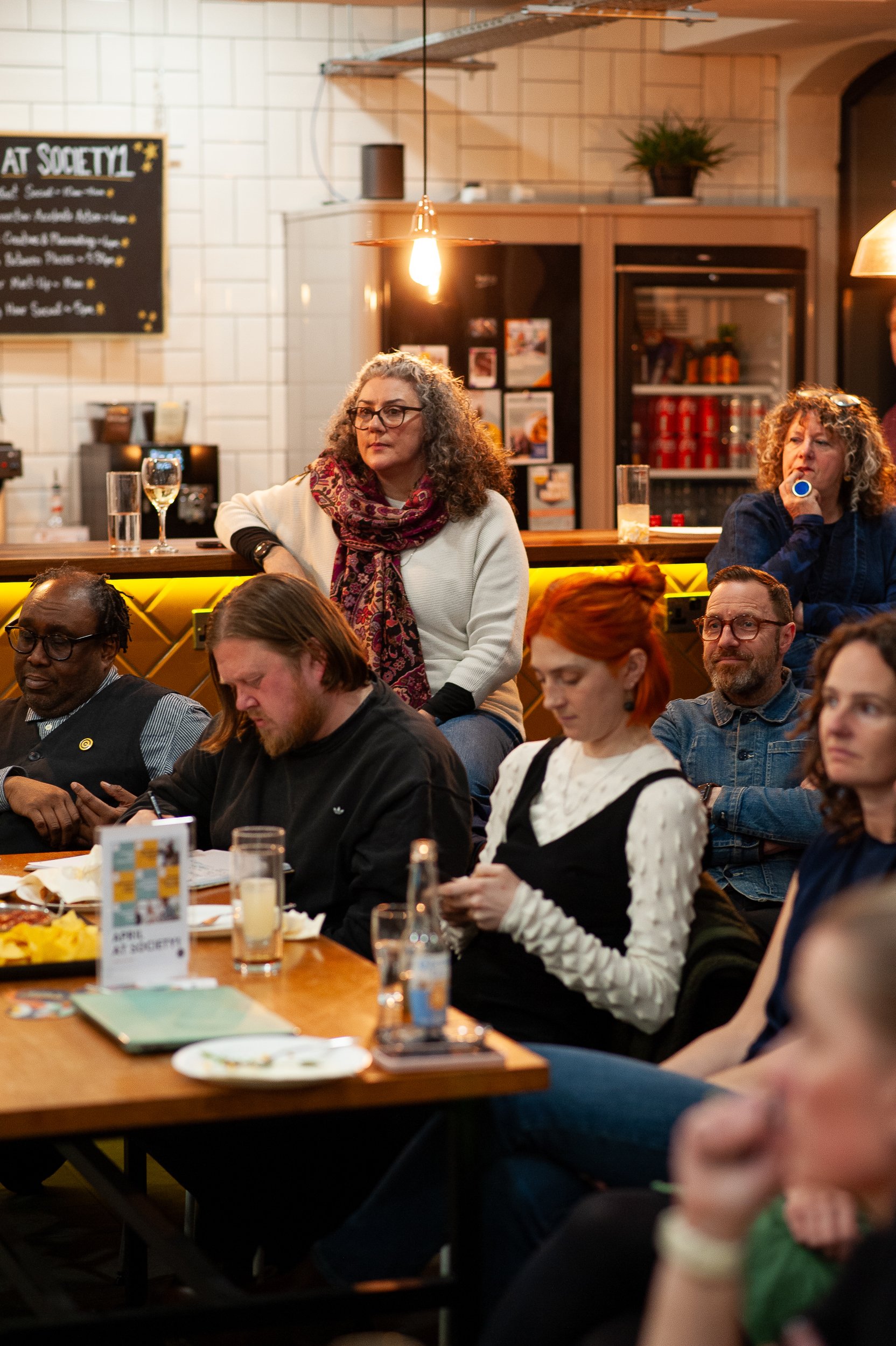 Group of people sitting in a cafe attending an event, some writing and others watching attentively. There's a menu board on the wall and drinks on the tables.