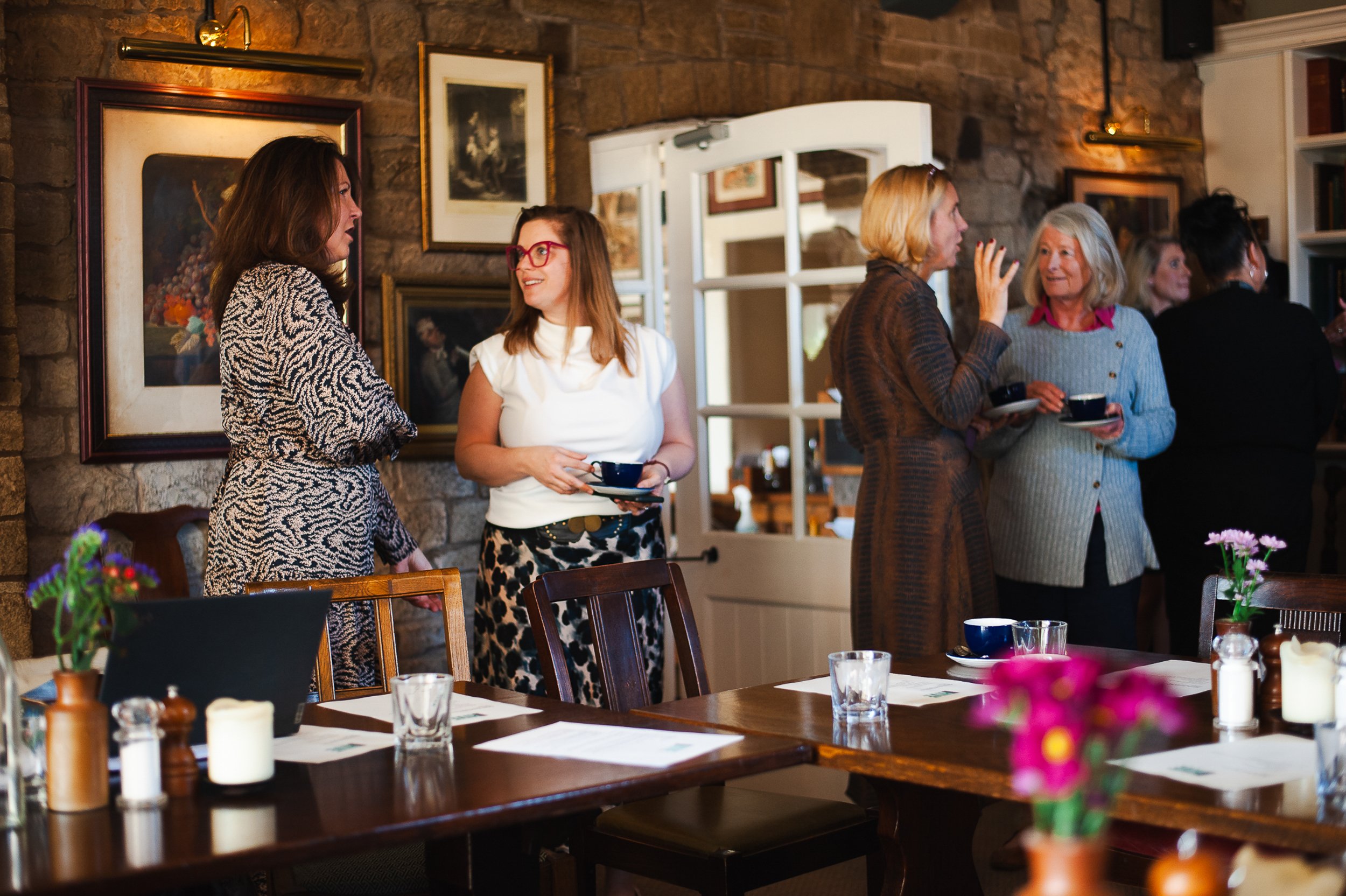 Group of women in a cozy restaurant setting, engaged in conversation and holding coffee cups, surrounded by framed artwork and wooden tables with flowers and glasses.