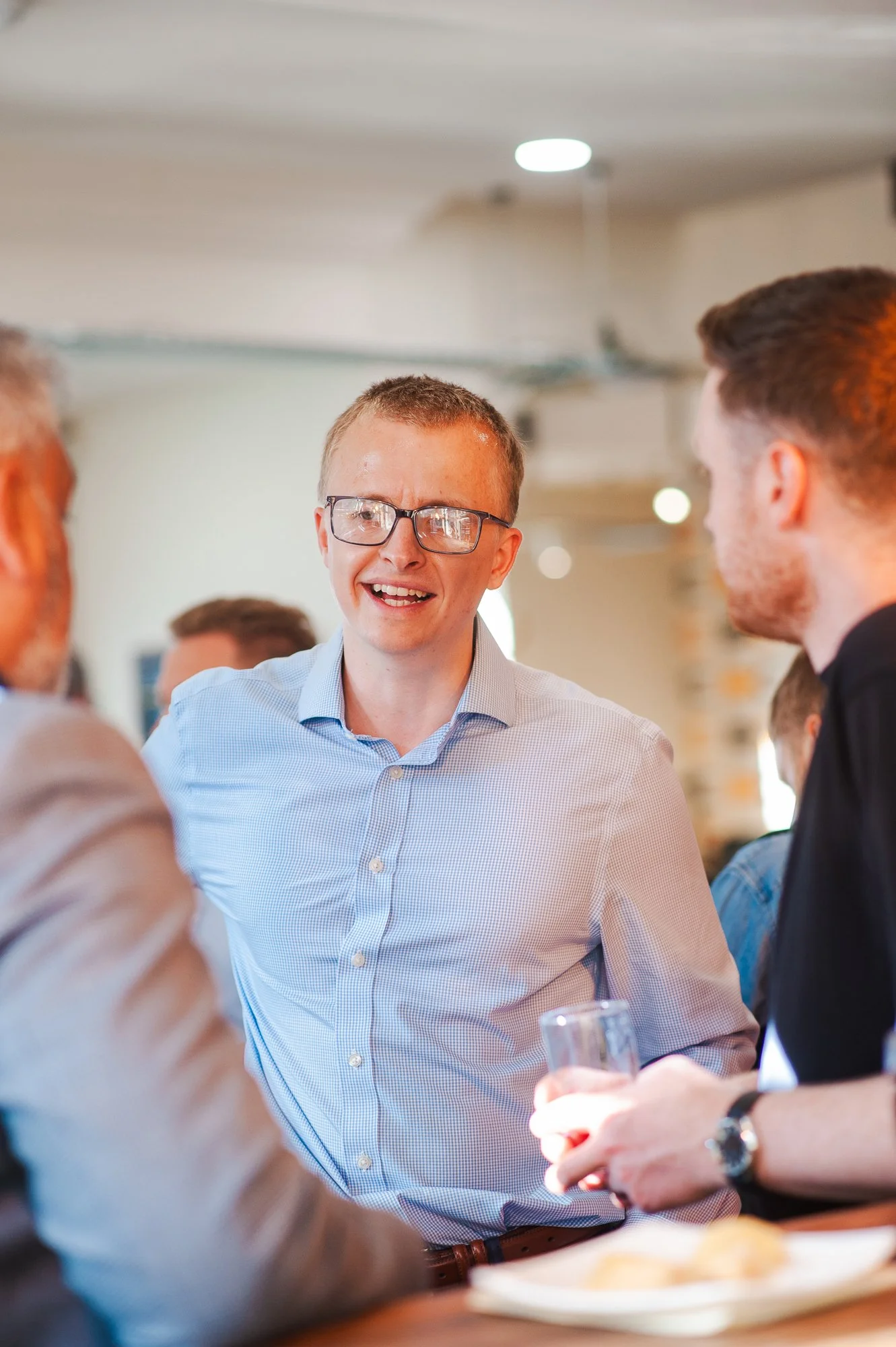 A young man with glasses and a blue checkered shirt smiling while talking to others in a social setting.