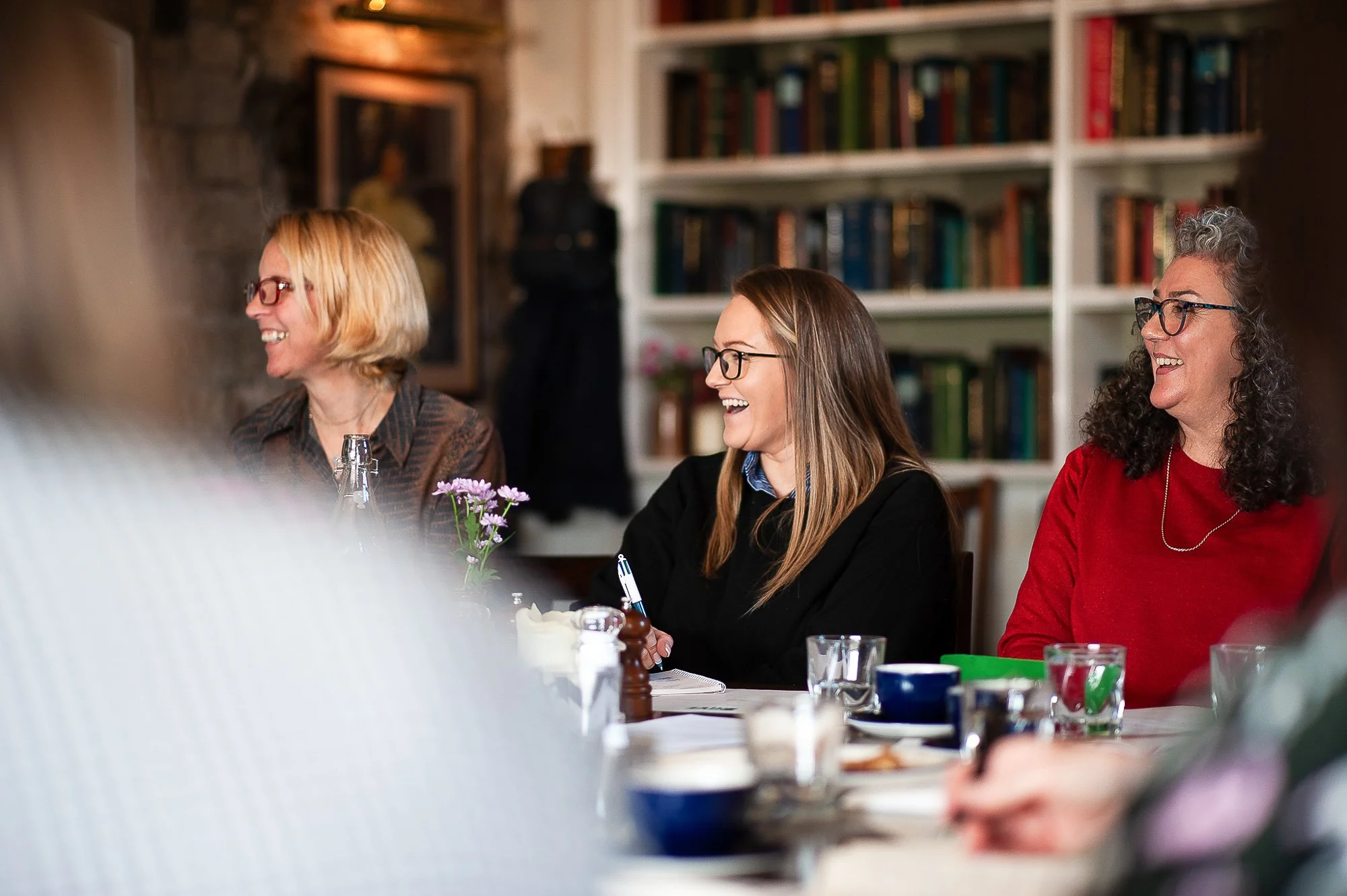 Three women seated at a table, smiling and engaged in conversation, with a bookshelf in the background.