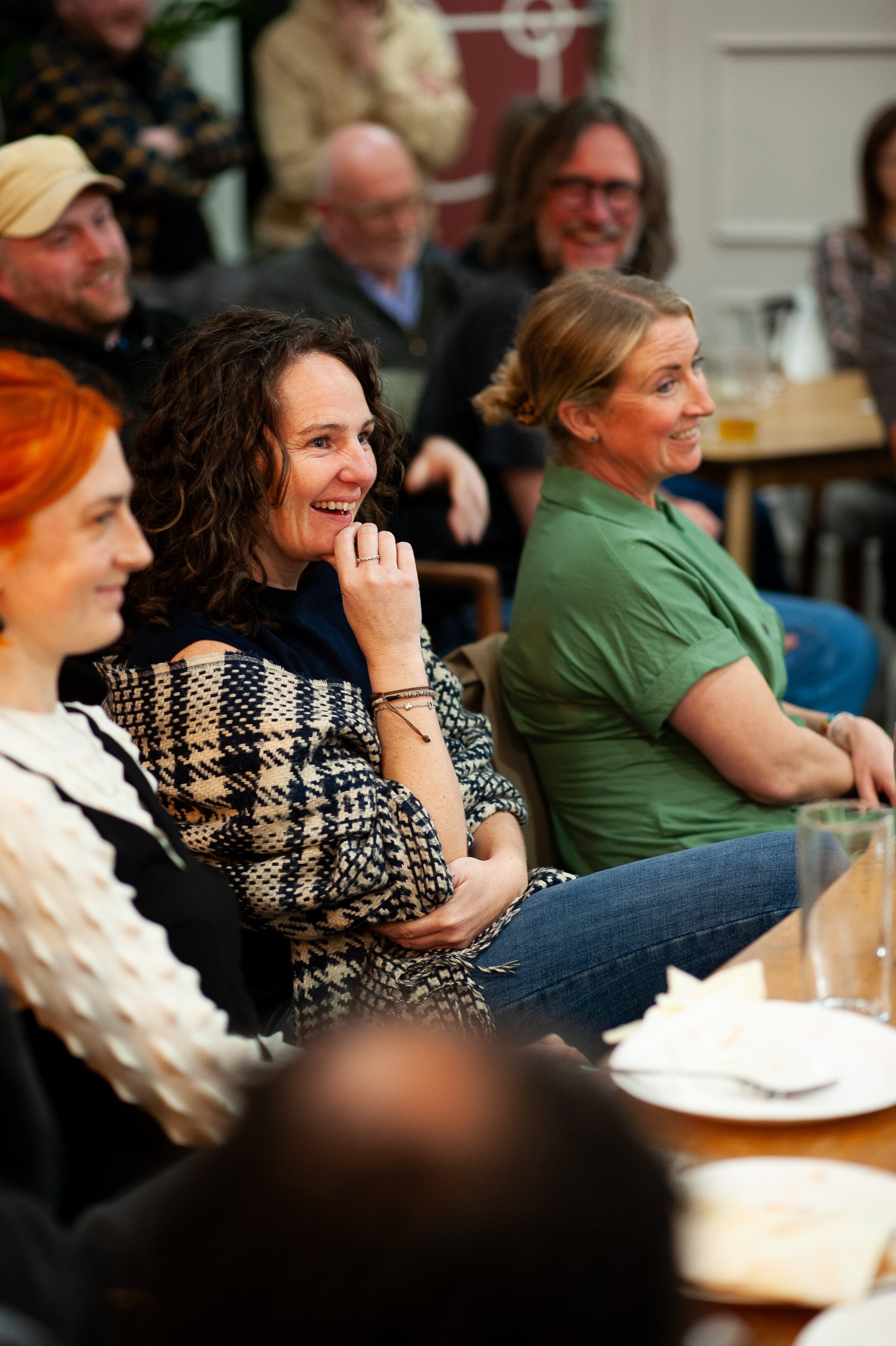 A group of people sitting in a room, smiling and watching something off-camera. The focus is on a woman in a checkered shawl, who is laughing. They seem to be enjoying a performance or presentation. Tables with glasses and plates are visible.