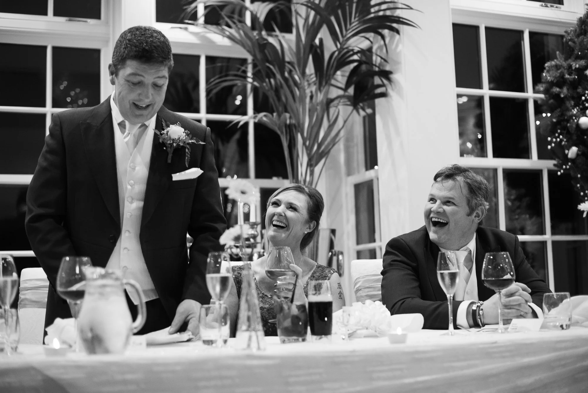 Three people celebrating at a dinner table, two men and one woman, smiling and enjoying drinks, in an elegant setting with large windows and plants in the background.