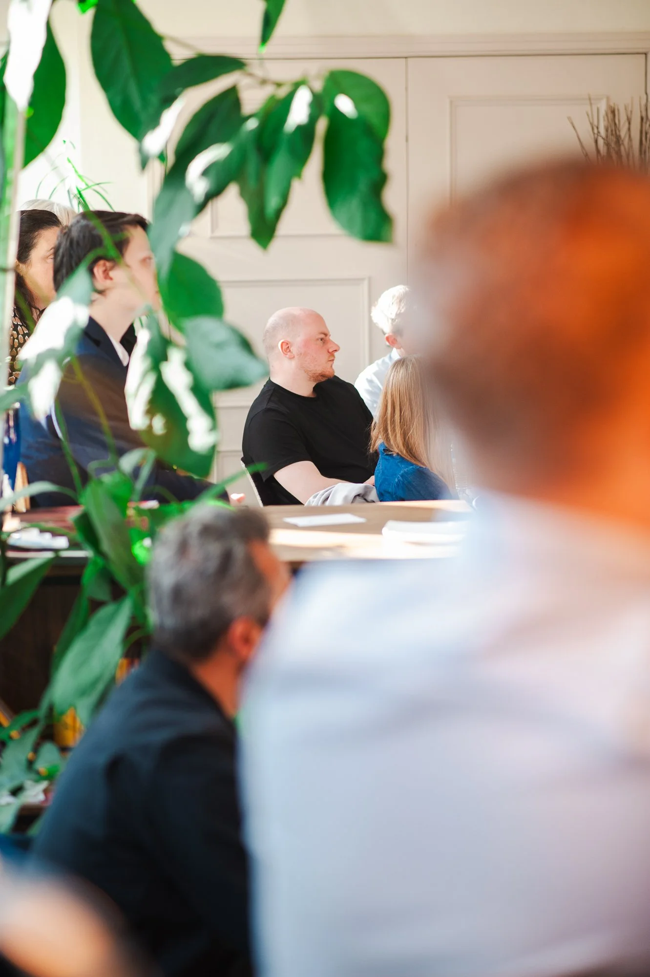 People attending a meeting or presentation sitting in a conference room, partially obscured by large green leaves in the foreground.