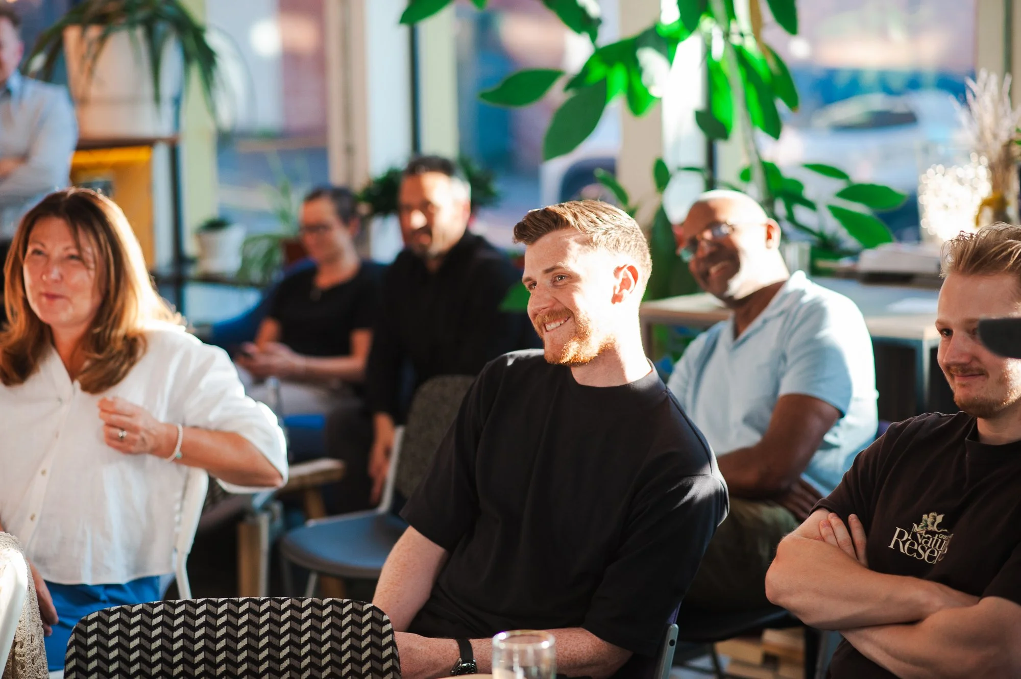 People listening to a speaker or presentation in a well-lit, cozy indoor space with plants and large windows.