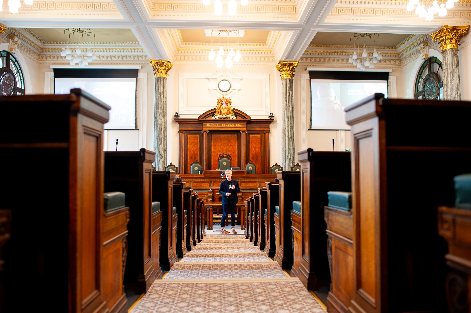 A man standing in a large, ornate courtroom with wood-paneled walls, chandeliers, and two large screens on either side. The courtroom has rows of wooden benches and columns with decorative gold accents.