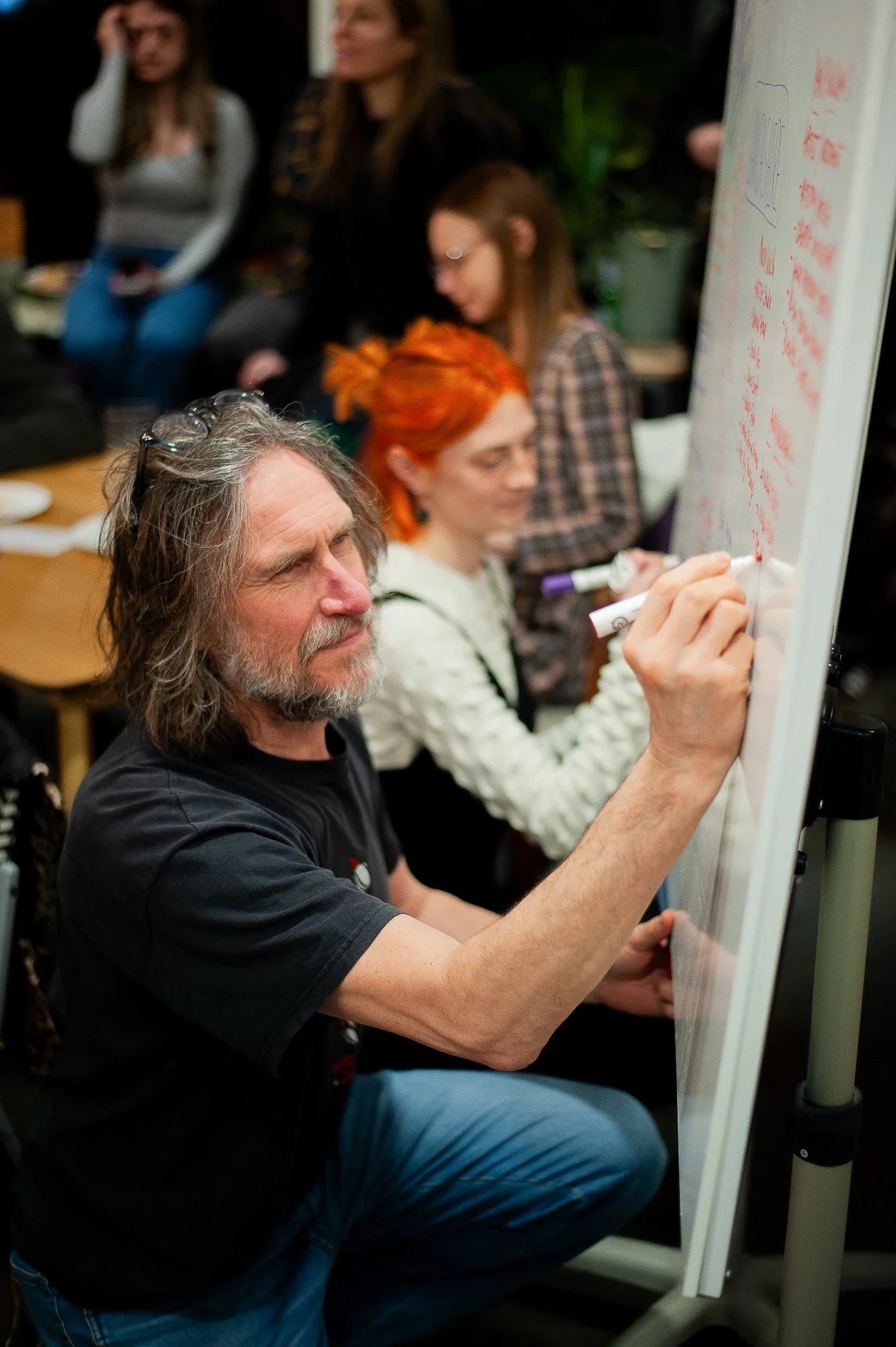 A man with gray hair and a beard writing on a whiteboard while seated, with several women, some with colorful hair, sitting and standing around in the background.