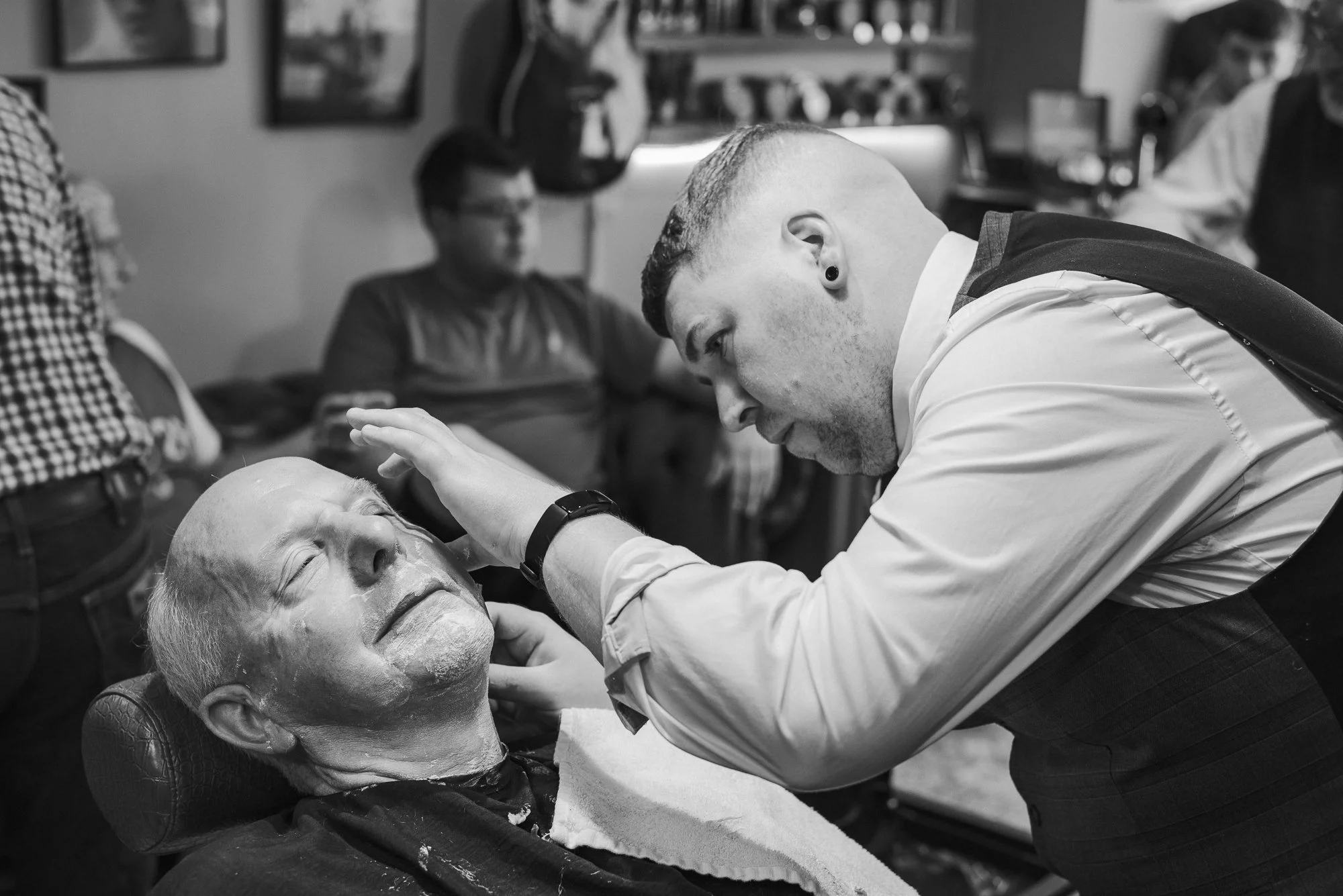 A man getting a beard shave in barbershop with barber leaning over him, while other customers sit in the background.