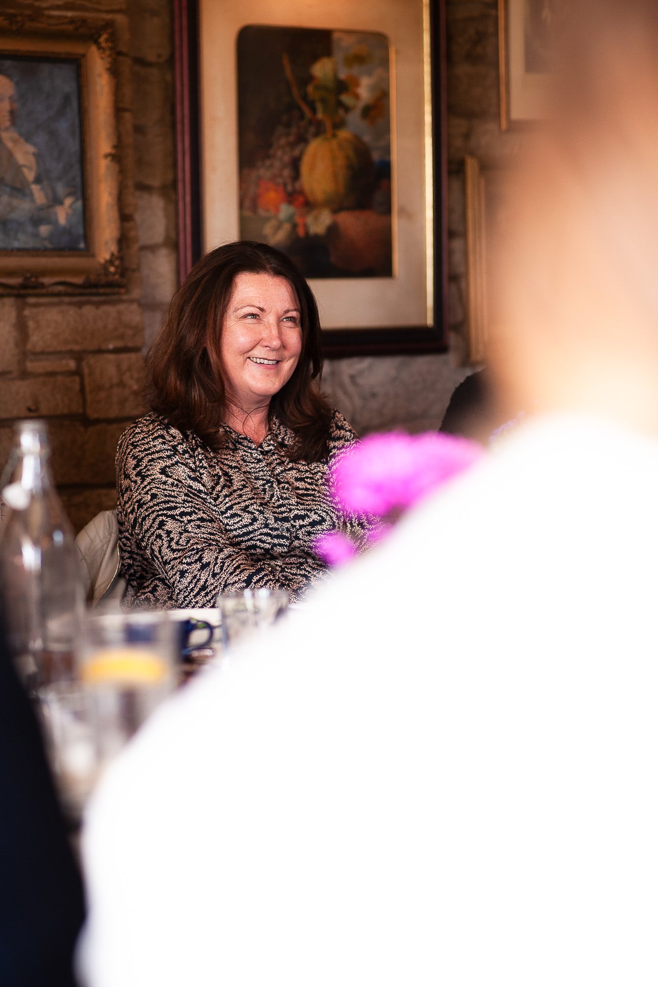 A smiling woman with brown hair sitting at a table during a social gathering or dinner. The background includes framed paintings and a stone wall.