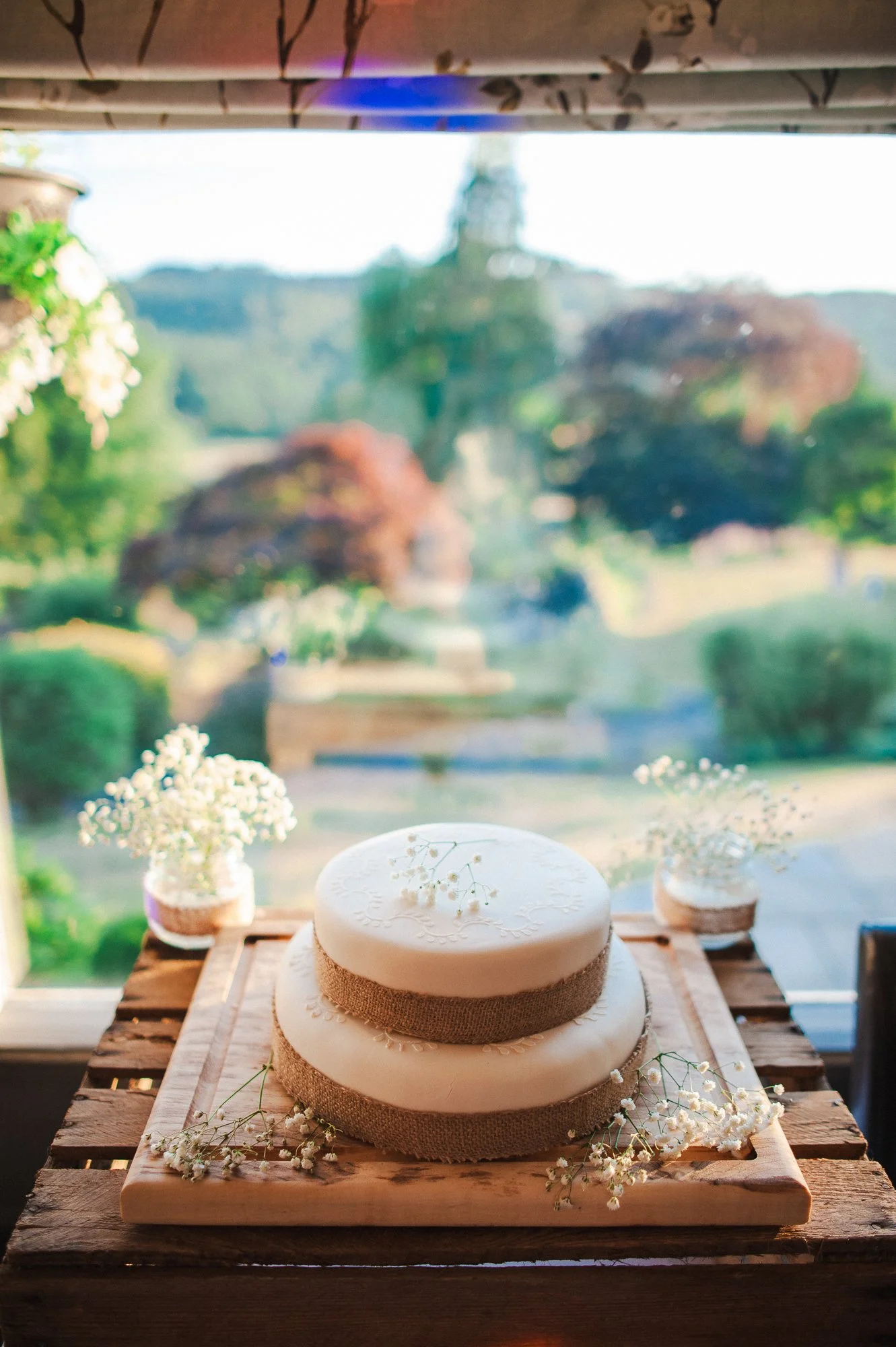 A two-tiered white wedding cake with burlap accents, decorated with small white flowers, on a wooden table near a window with a scenic outdoor view.