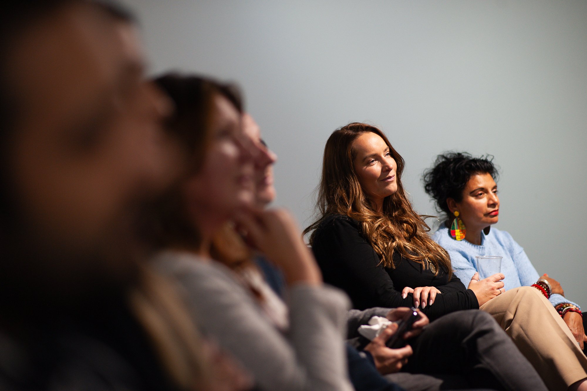 Four women sitting in a row, attentively listening during a discussion or presentation, with one woman holding a glass of water and wearing colorful earrings.