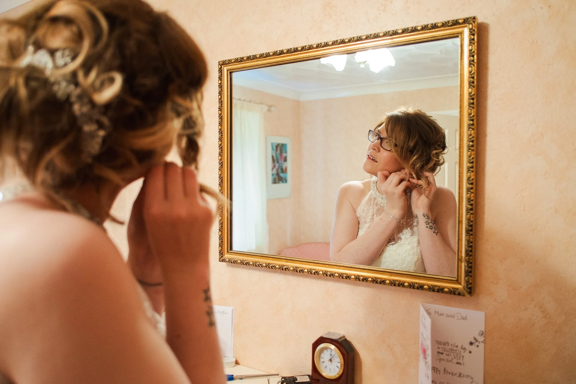 A woman with glasses, wearing a white lace dress, looks at herself in a wall mirror while adjusting her hair, preparing for a special occasion.