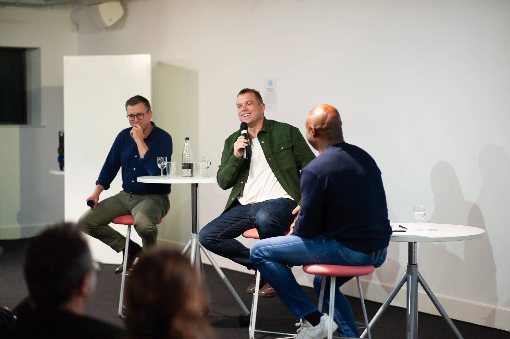 Three men sit on high stools on a stage in front of an audience, participating in a panel discussion. The man in the middle is speaking into a microphone, smiling, and wearing a green jacket and white shirt. The man on the left is wearing glasses, a 