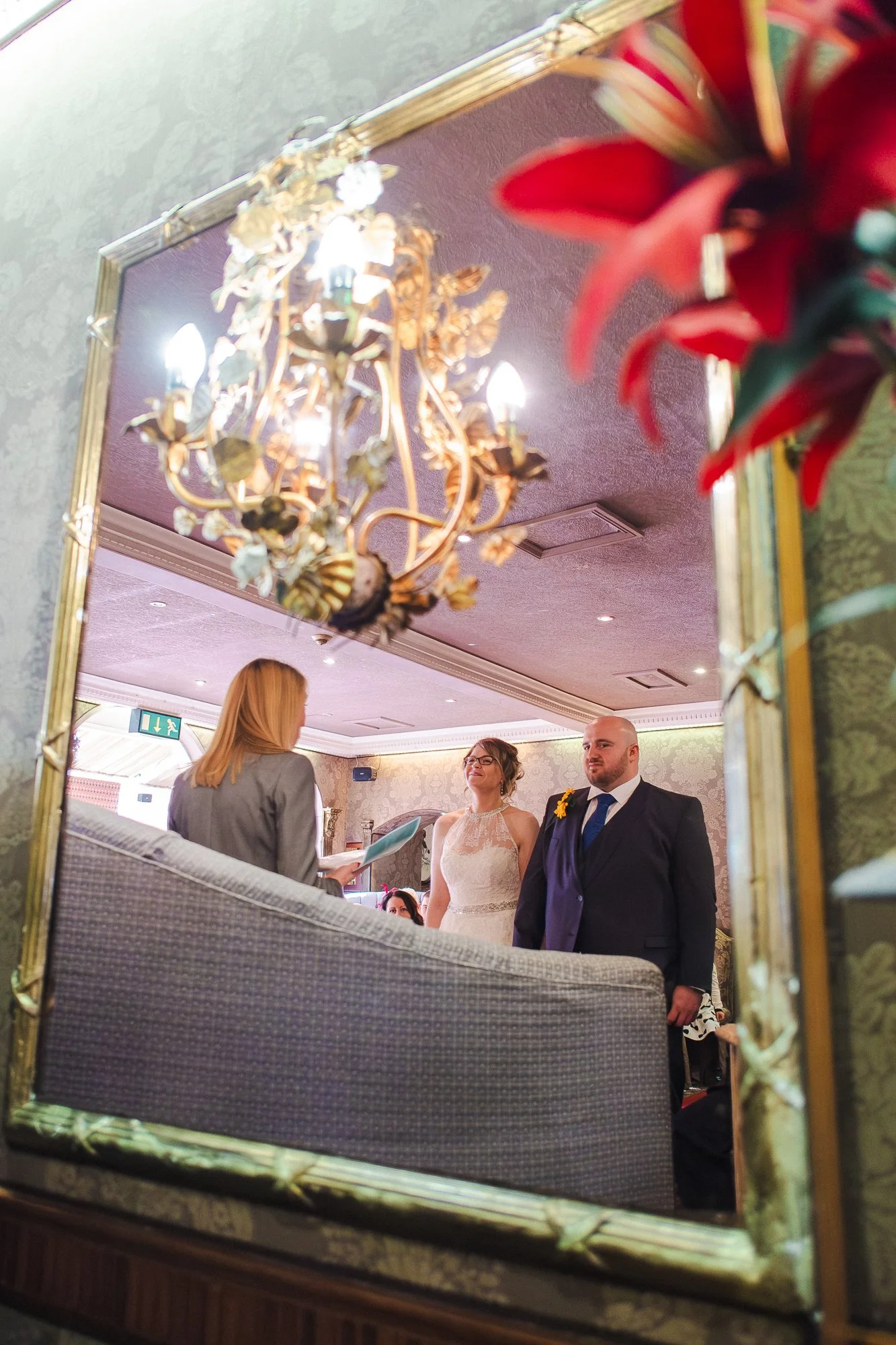 Bride and groom standing during their wedding ceremony, reflected in a mirror with a gold frame, in a decorated indoor venue with a chandelier and floral arrangements.