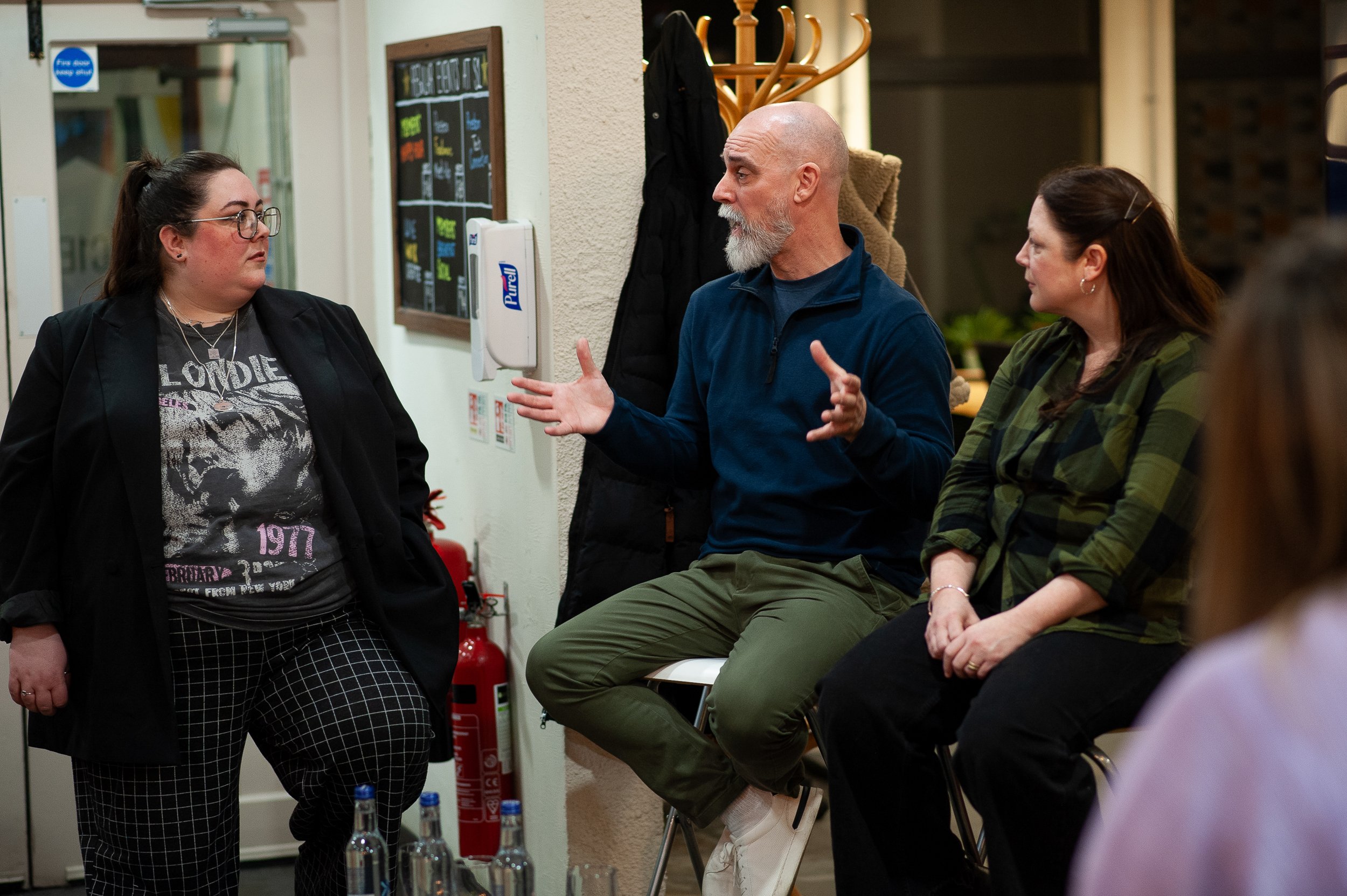 Three people having a discussion indoors; one standing and two seated on stools, with a coat rack and a wallboard in the background.