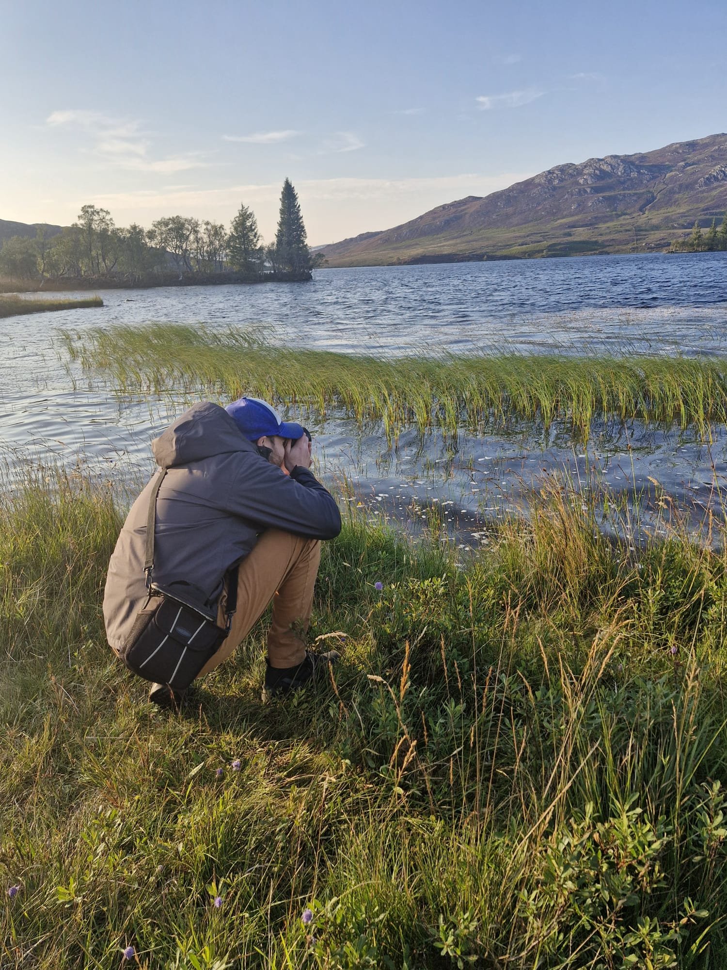 Person crouching on grassy lakeshore, taking a photograph of the lake and mountains in the distance during daylight.