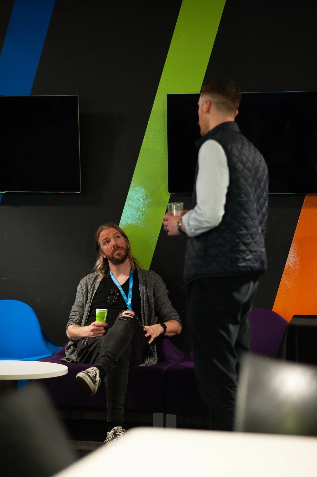 Two men having a conversation in a modern lounge with colorful wall panels and wall-mounted screens; one man is sitting on a purple couch, holding a drink, looking up at the other who is standing, holding a glass, with a coffee cup on the table nearb