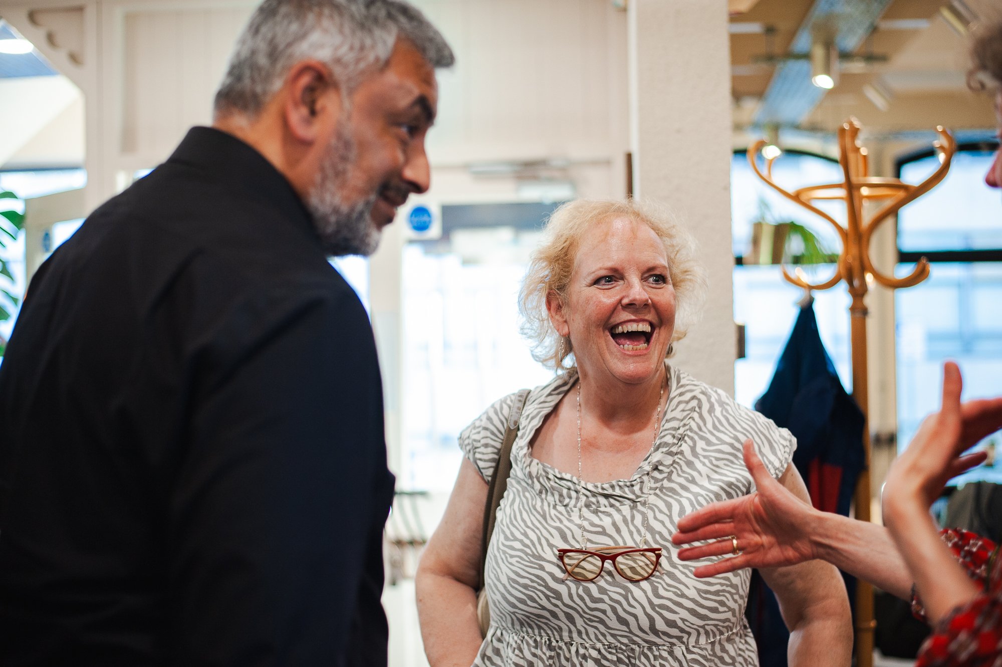 Three people having a lively conversation, smiling and laughing indoors. One woman with curly blonde hair and eyeglasses hanging on her shirt is speaking to a man with gray hair and a beard, and a person with hand gestures partially visible.