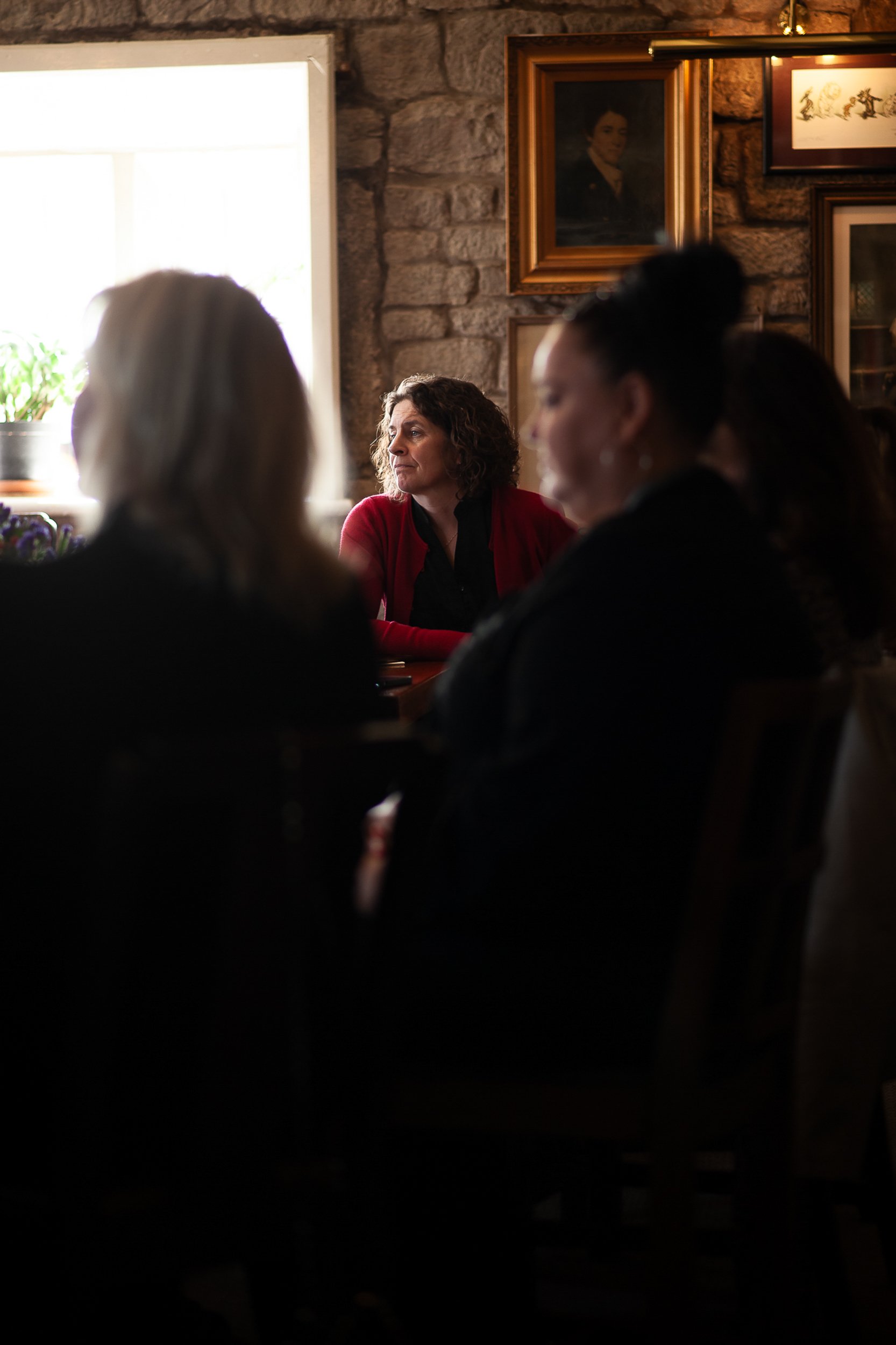 Group of people seated indoors; a woman in a red cardigan looking out a window.