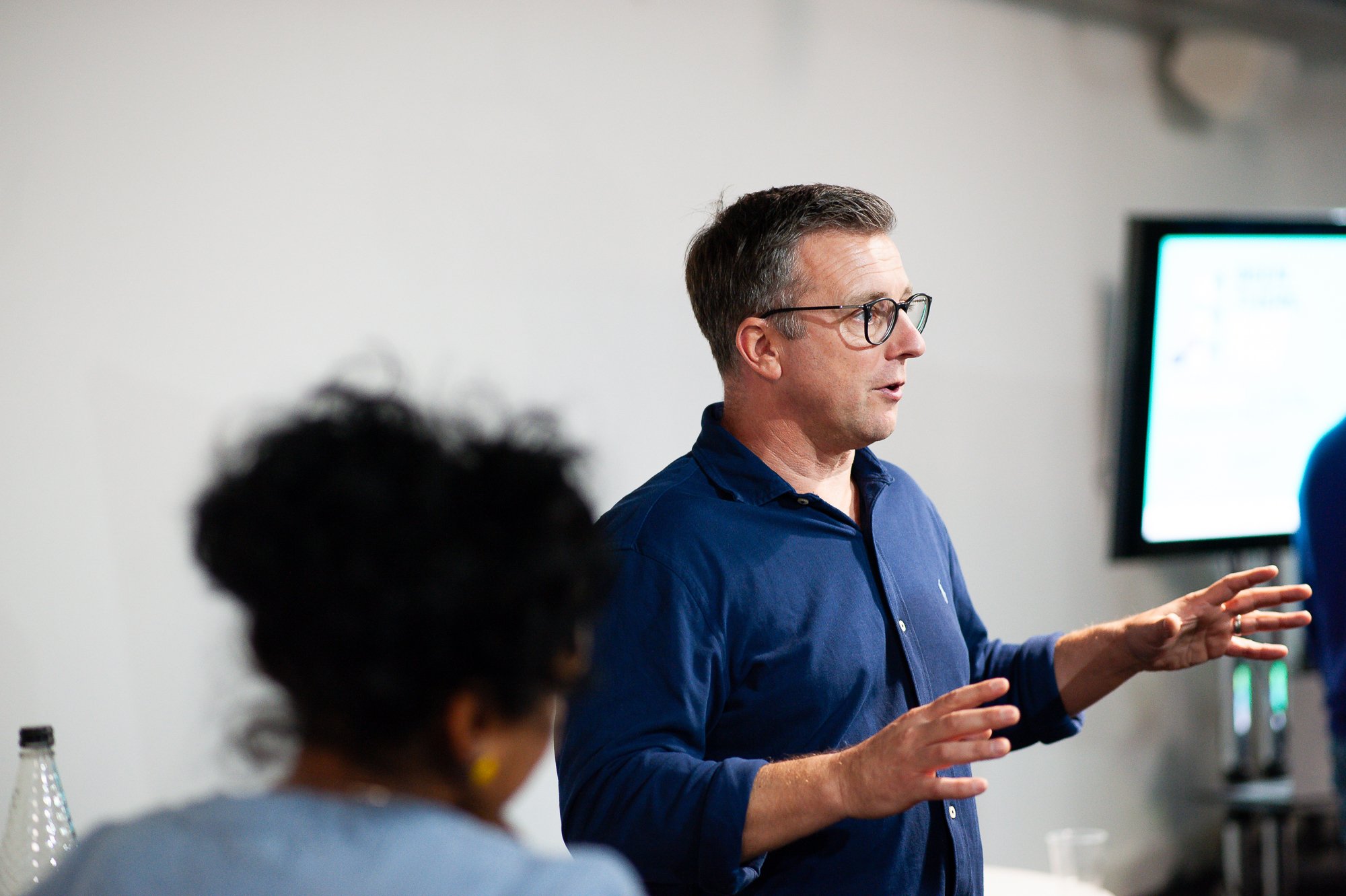 A man with glasses and short dark hair, wearing a blue button-up shirt, is speaking or presenting in front of a room with people and a large screen in the background.