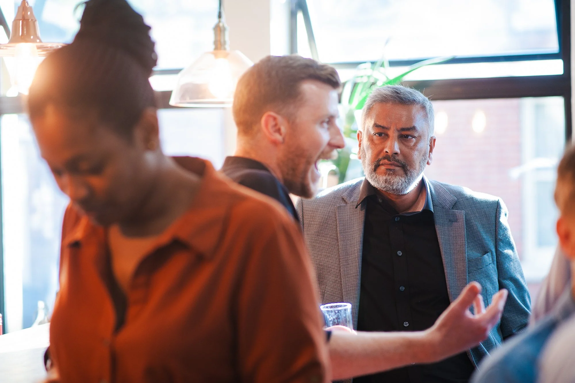 Group of four people having a conversation in a coffee shop, with natural light coming through large windows behind them.