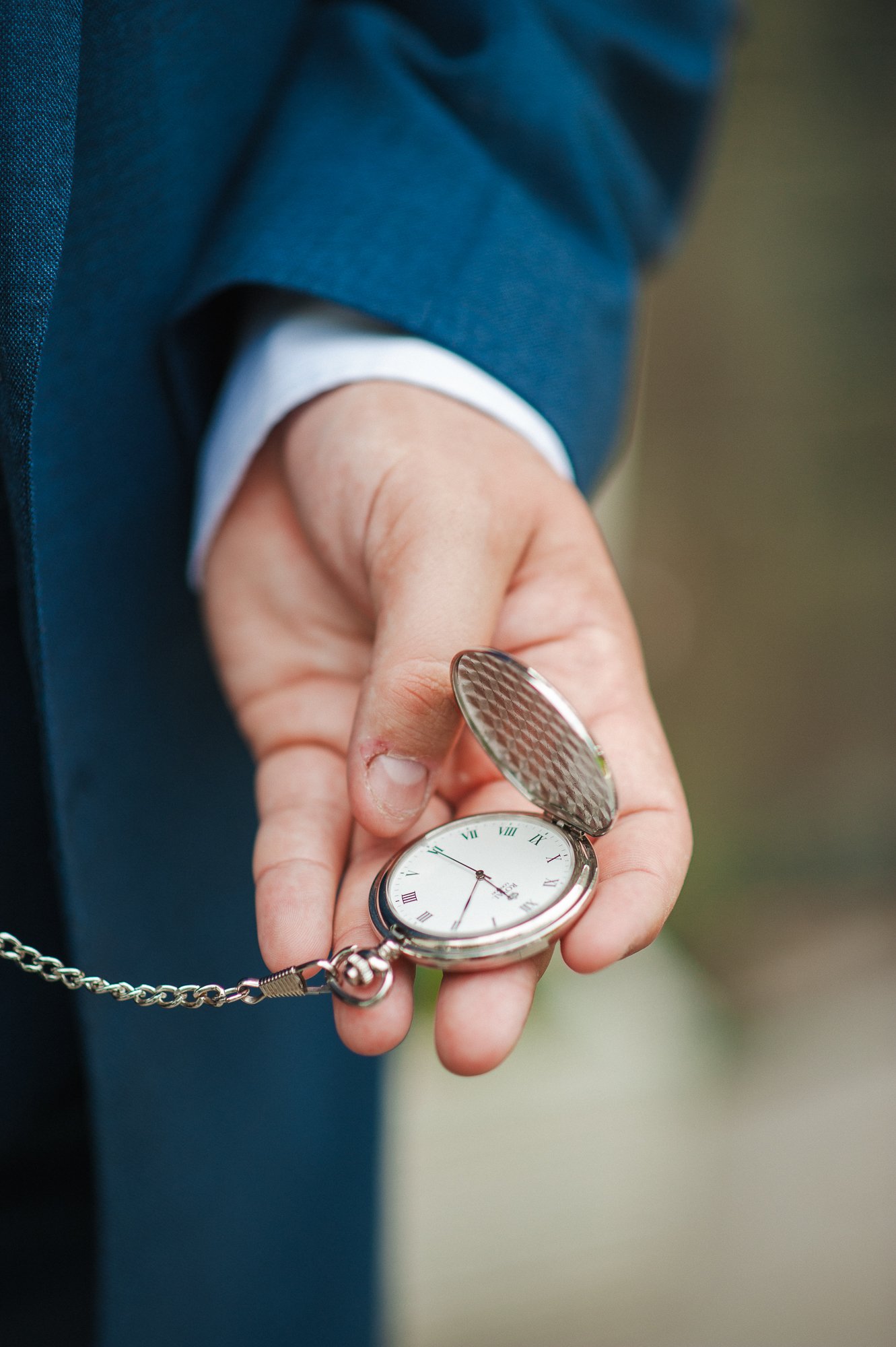 A person in a suit holding a silver pocket watch with a chain, with the watch's cover open showing the clock face.