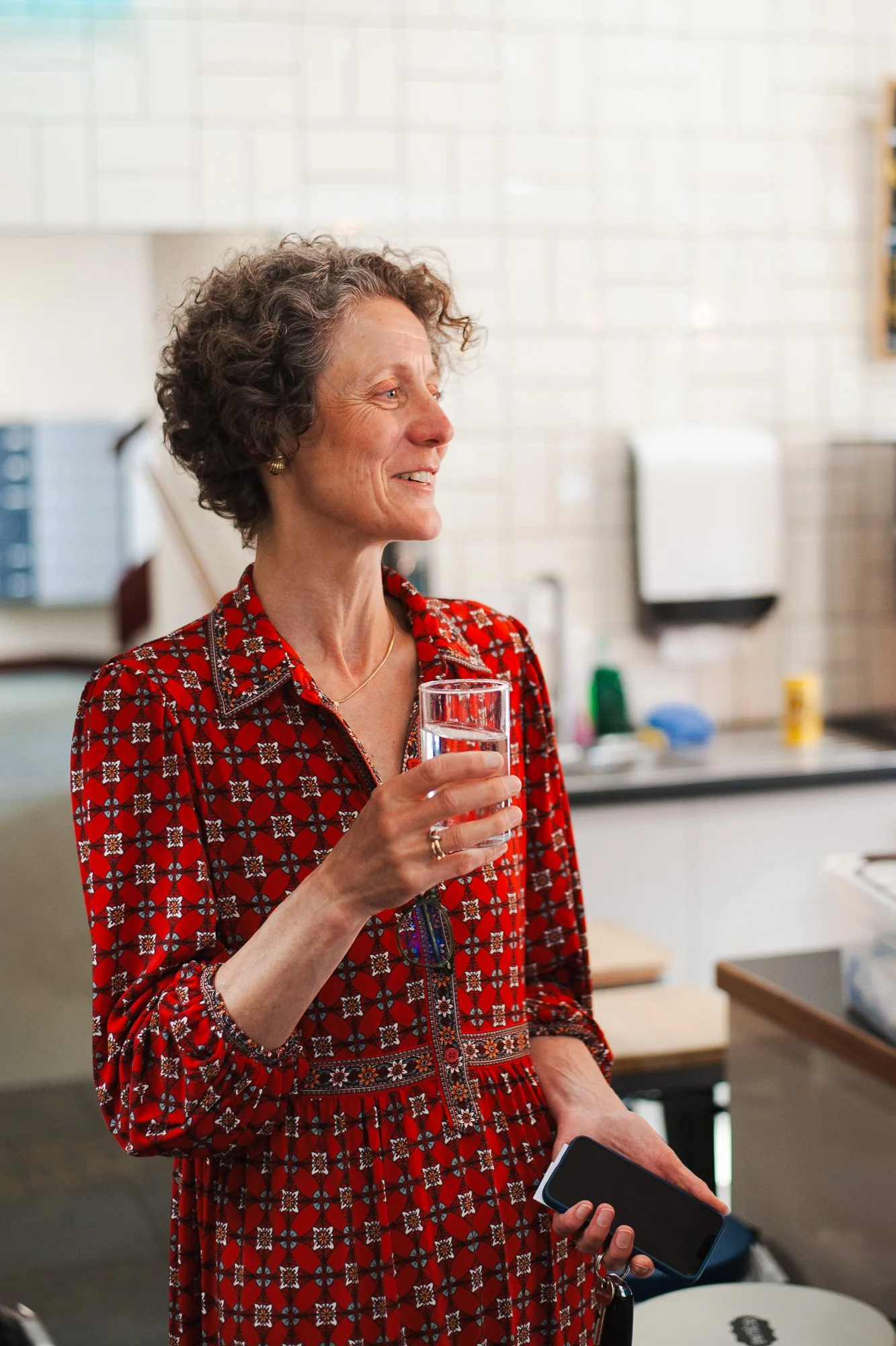 A woman with curly gray hair wearing a red patterned dress is holding a glass of water and a smartphone, standing in a kitchen.