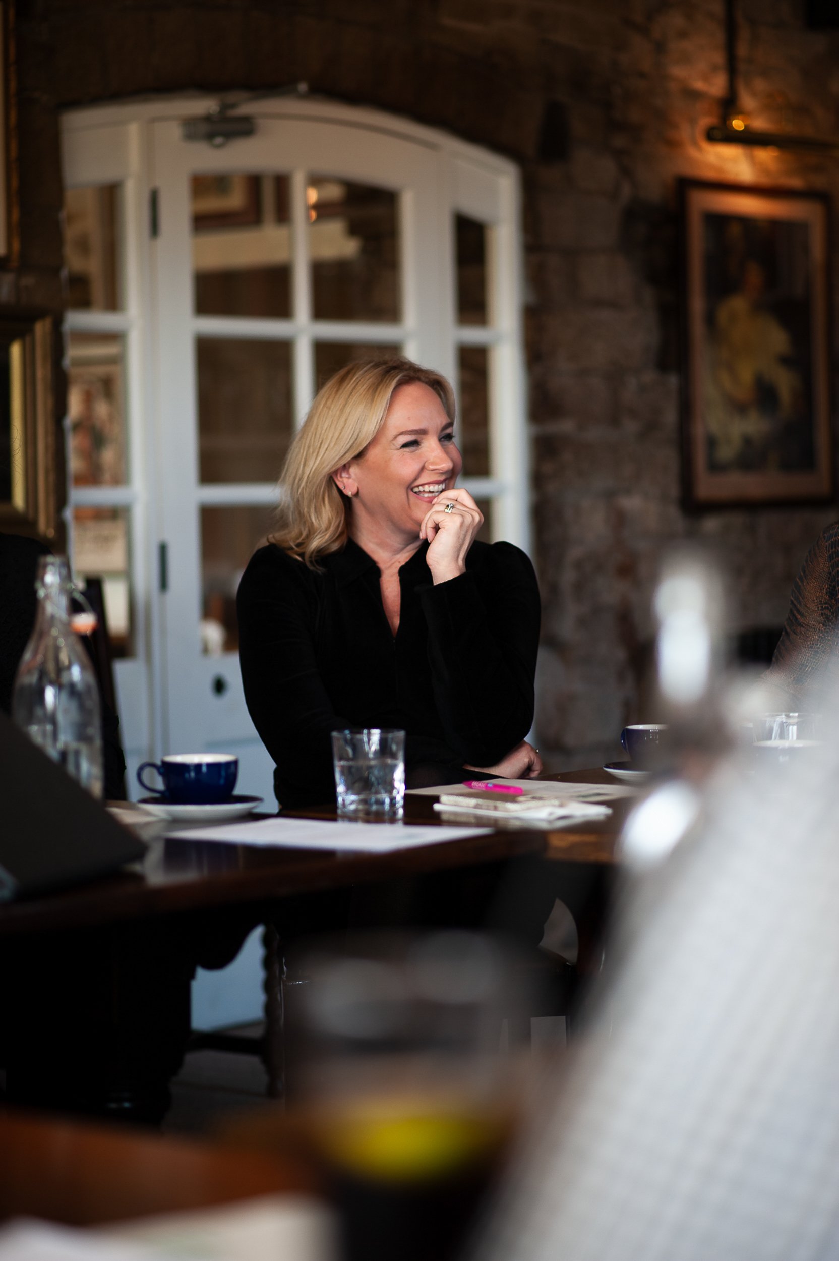 Blonde woman smiling at a table during a meeting, with cups and papers on the table.
