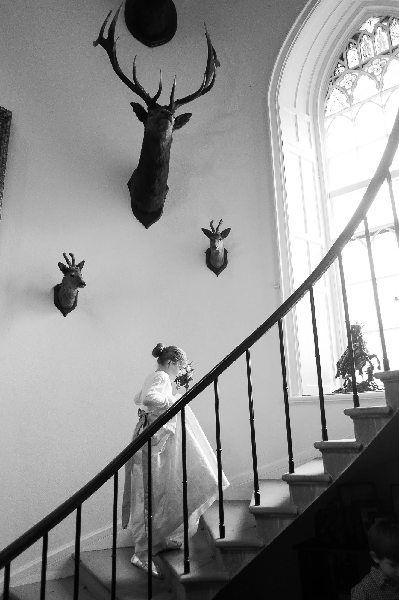 A girl in a dress walking up a staircase with deer head trophies mounted on the wall beside a large window.