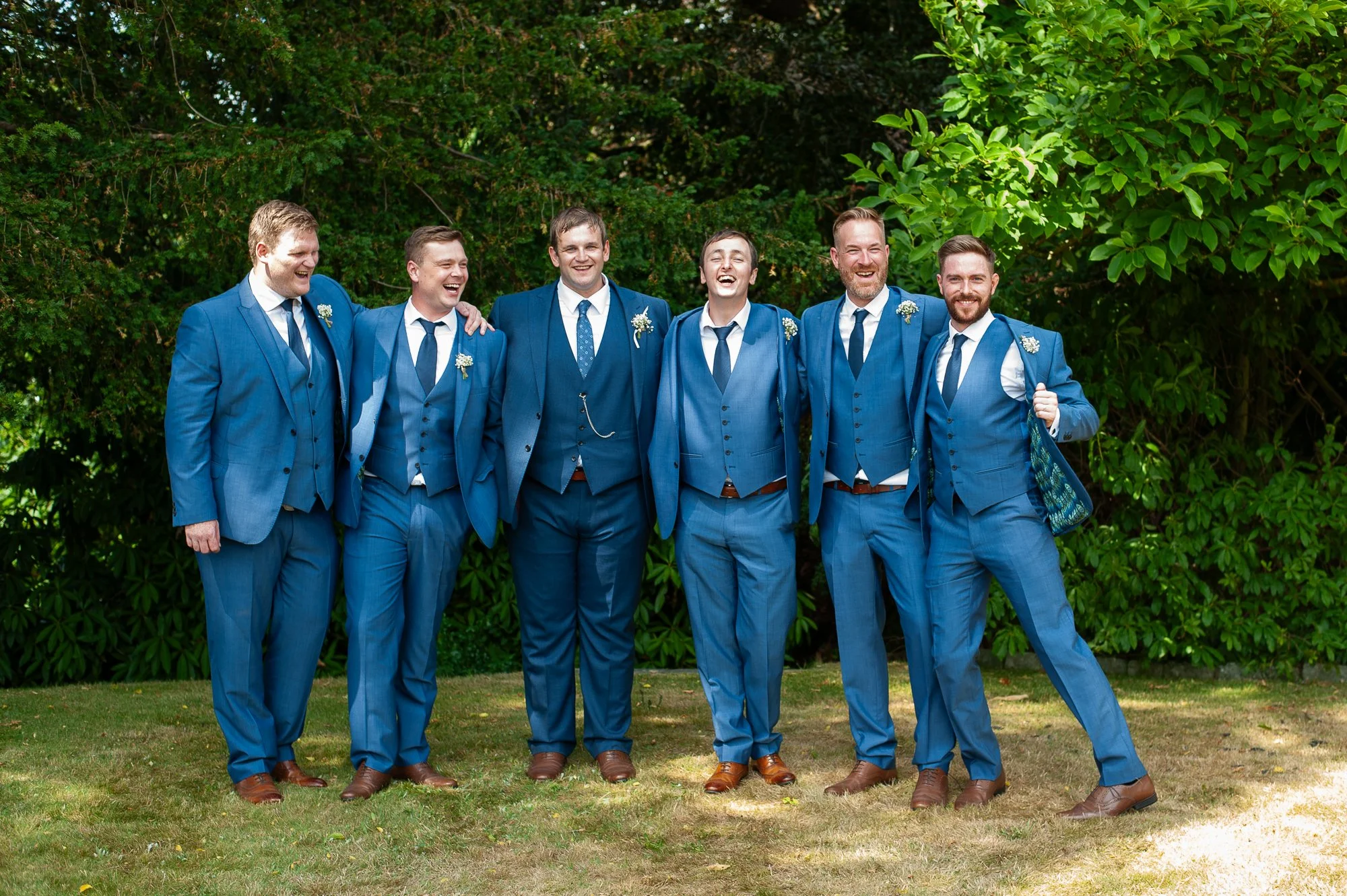 Seven men in blue suits enjoying outdoor gathering, standing on grass with green foliage and trees in background, some with arm around each other's shoulders, smiling and laughing.