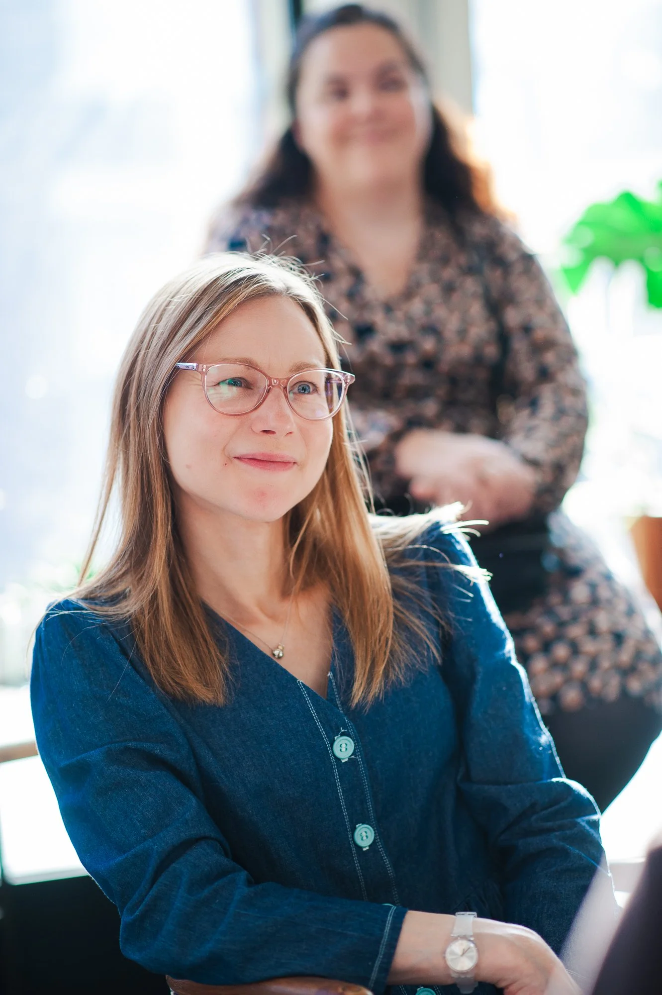 A woman with light brown hair, glasses, and a blue top sitting indoors with a smile, and a woman in the background with dark hair and a patterned blouse.