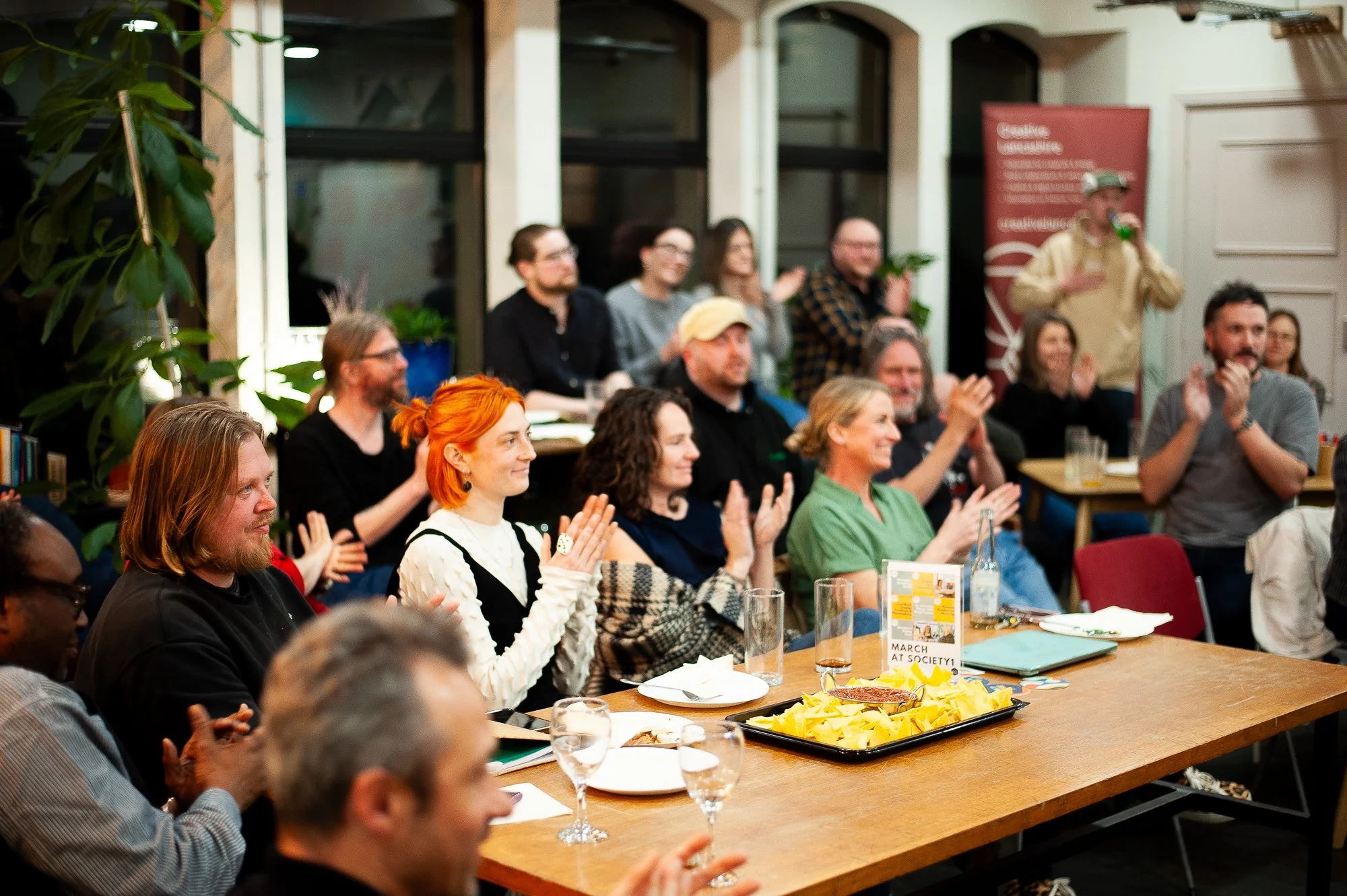 People gathered at a dinner or event, sitting at tables, clapping and smiling, with a person drinking from a green bottle in the background.