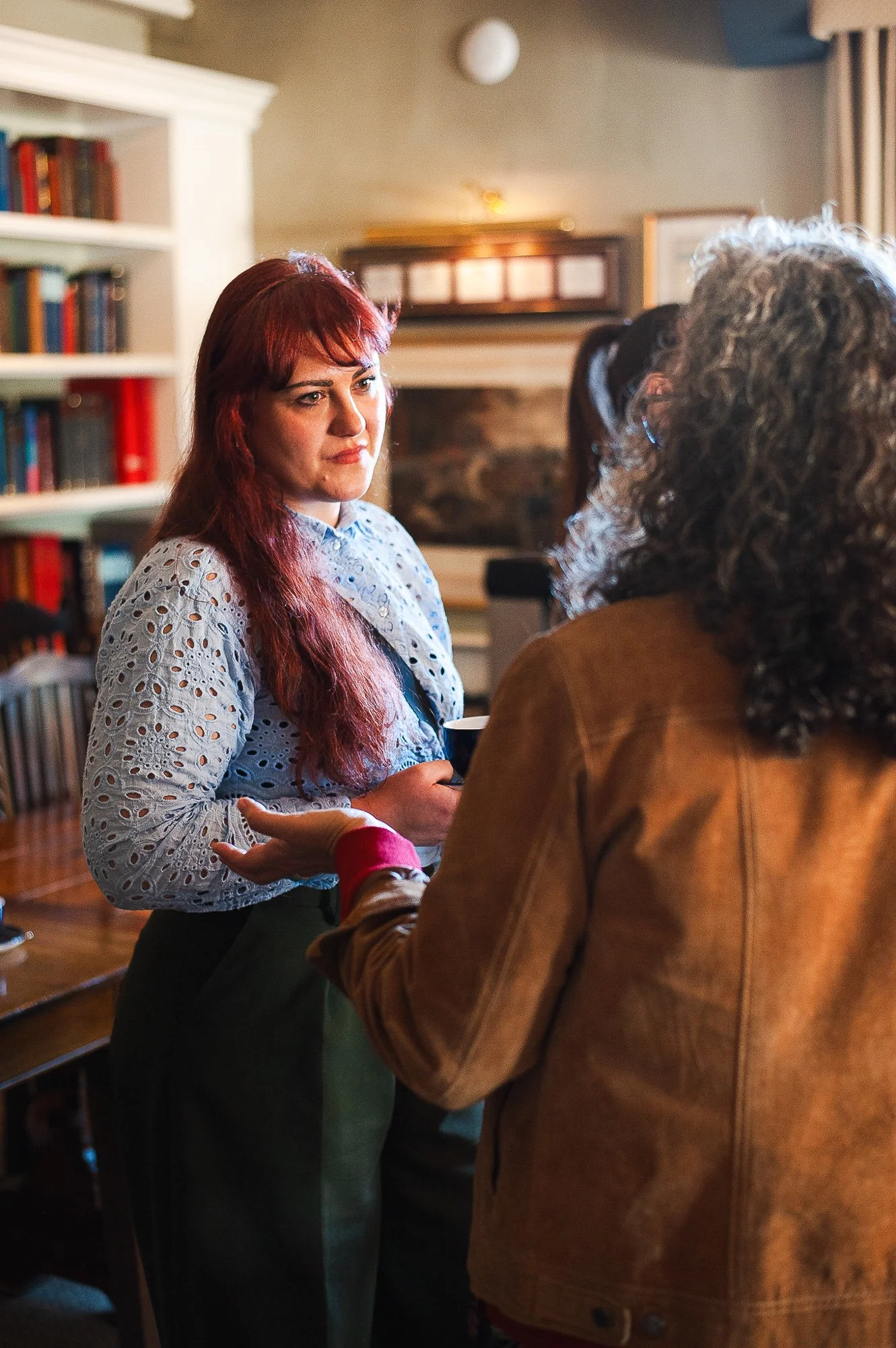 Two women are talking in a cozy room, one with red hair in a eyelet blue blouse holding a coffee mug, the other with gray curly hair and a brown jacket, engaging in conversation.