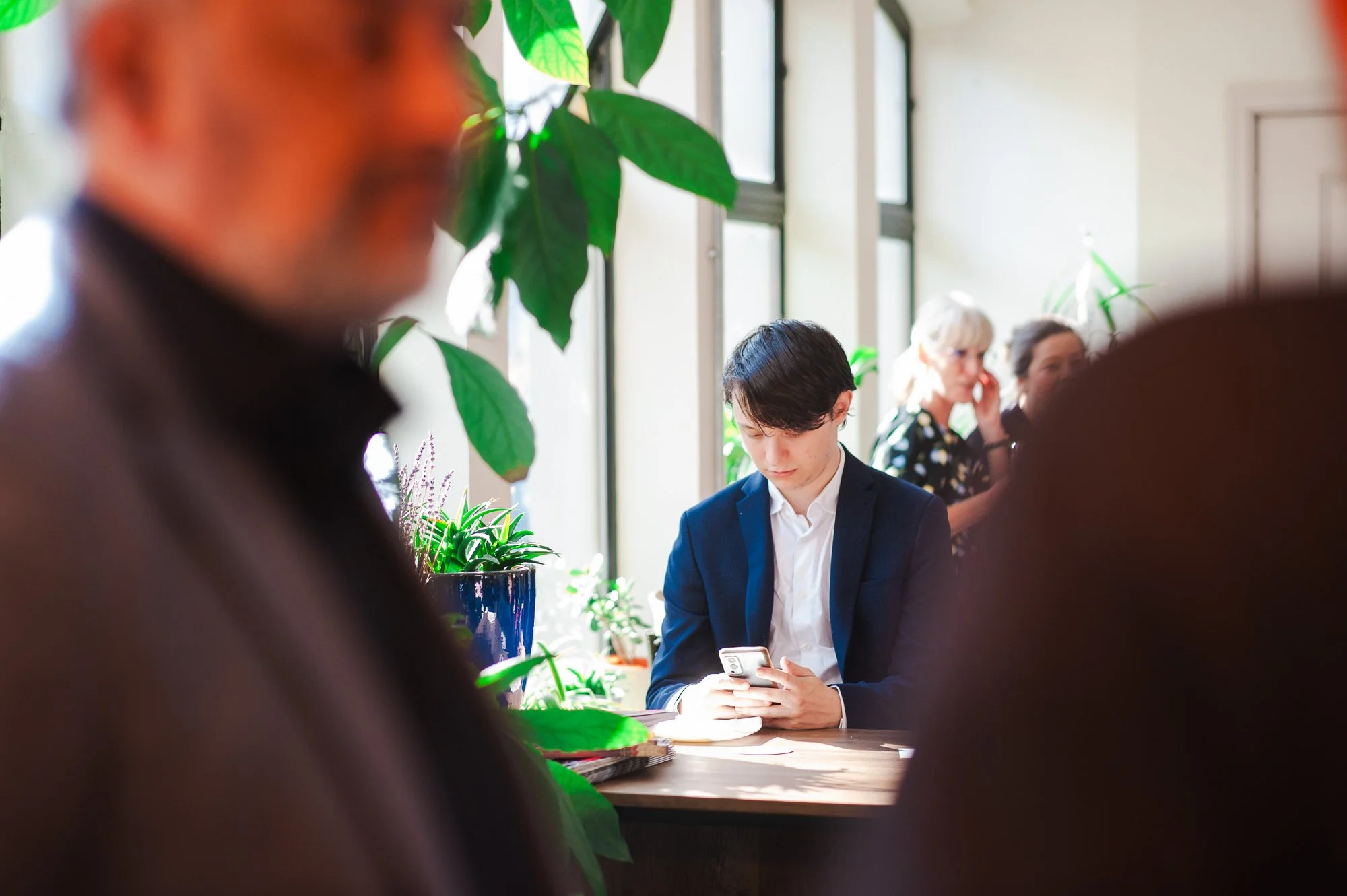 Young man in a suit using a smartphone at a table surrounded by people and green plants in a bright indoor setting.