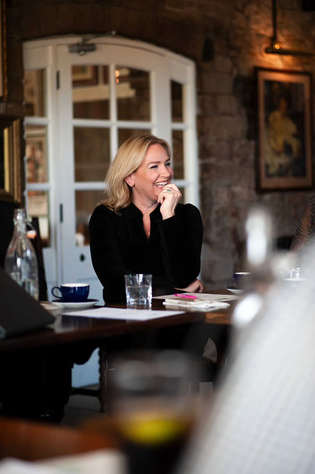 A woman sitting at a table, smiling and laughing during a meeting or gathering in a cozy, rustic room with stone walls and framed artwork.