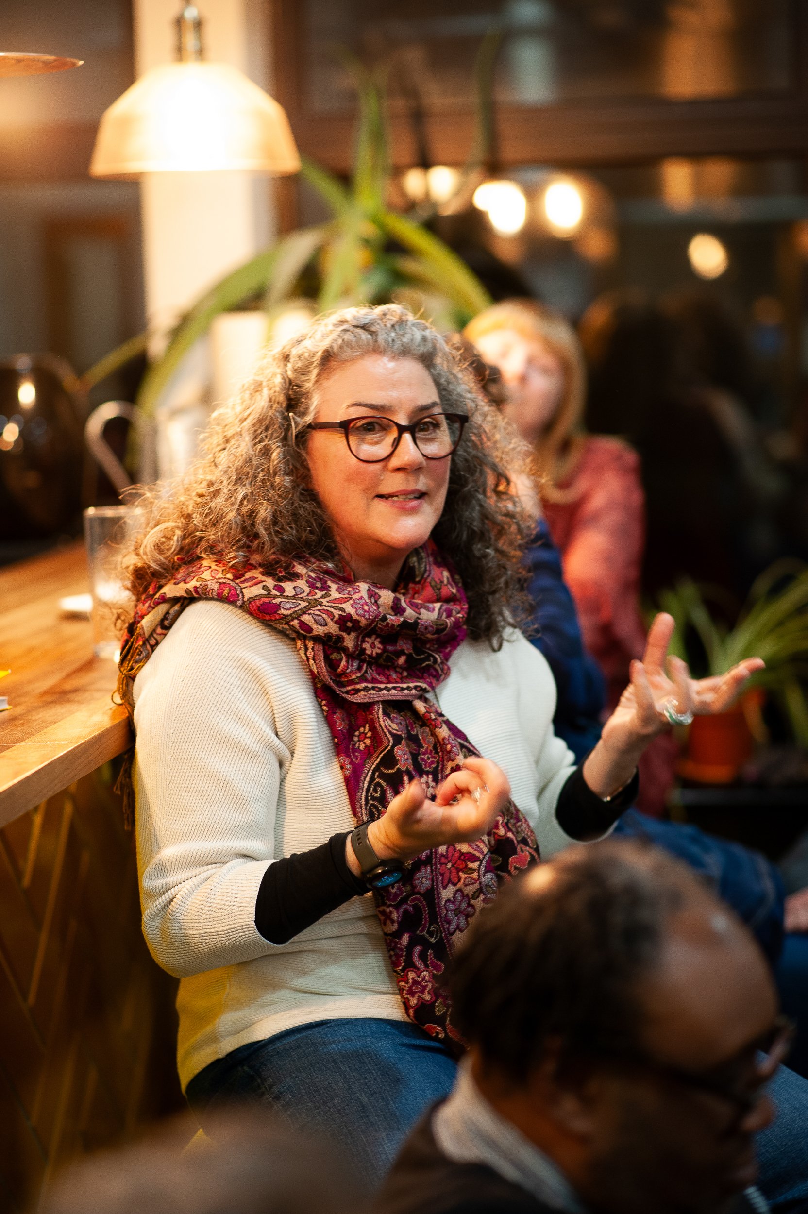 A person with curly hair and glasses, wearing a patterned scarf and light sweater, sitting indoors and gesturing while talking. Several people and indoor plants are visible in the background, with warm lighting.